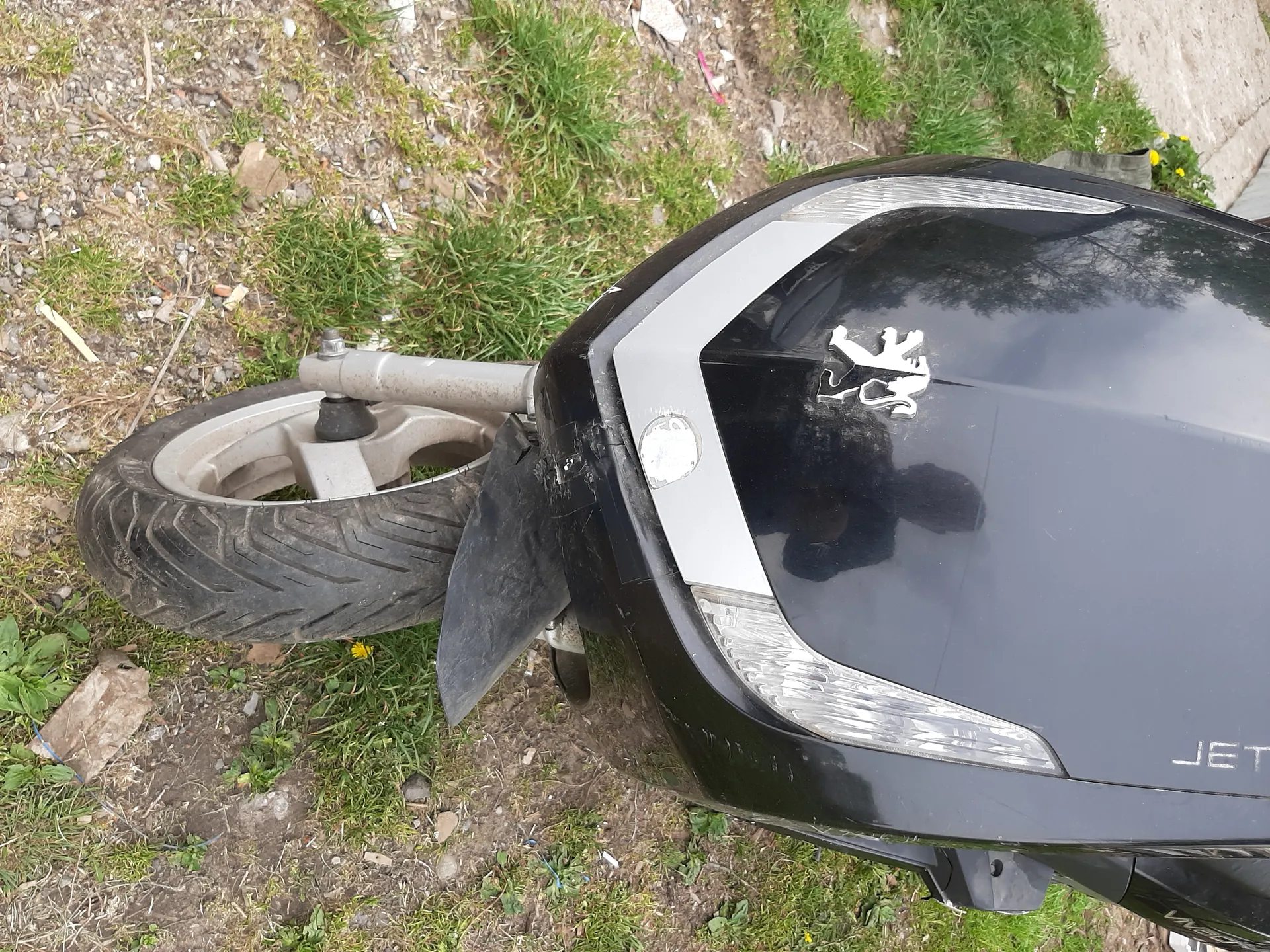 Czarny skuter marki Peugeot z widocznym białym logo lwa na nadwoziu. Widoczna część przodu skutera z reflektorem, kołem przednim oraz napisem JET na nadwoziu. Tło stanowi trawa, ziemia i rozrzucone drobne przedmioty.