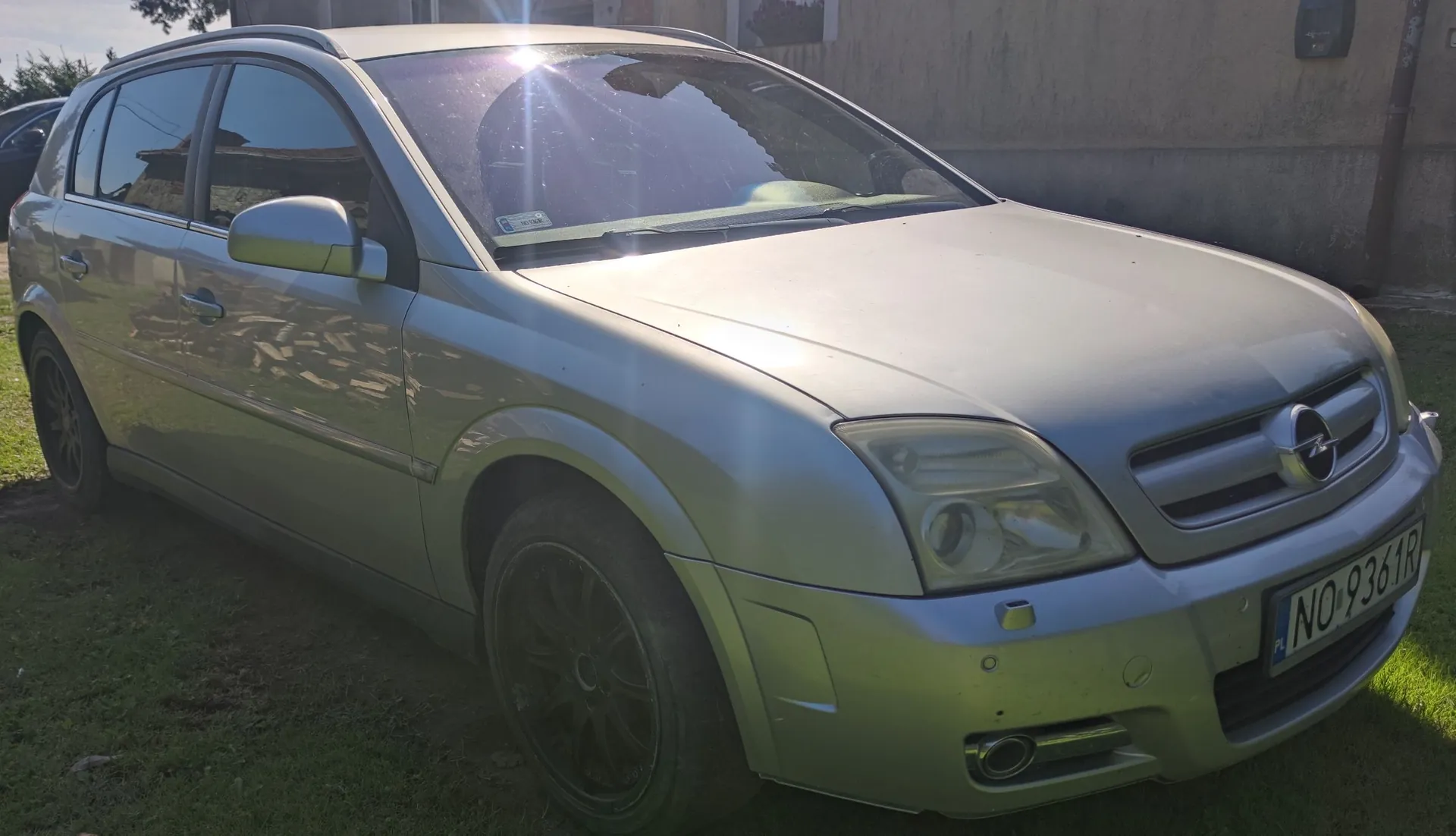 Srebrny samochód marki Opel w nadwoziu kombi, zaparkowany na trawie przed budynkiem. Widoczny jest przód i lewy bok pojazdu. Samochód ma tablicę rejestracyjną o numerze NO 9361R.