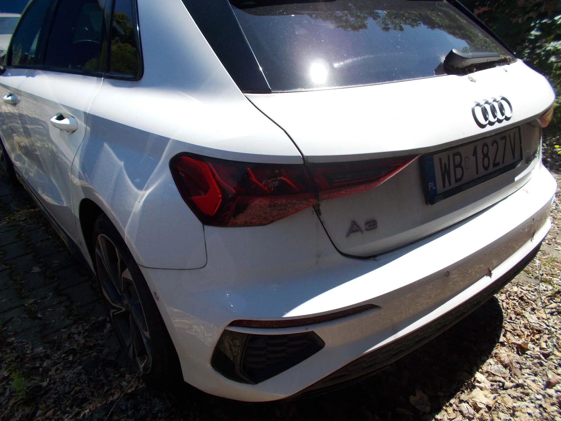 Tylna część białego samochodu marki Audi A3. Widoczny jest emblemat czterech pierścieni, oznaczenie modelu 'A3' oraz tablica rejestracyjna o numerze WB 1827V. Samochód zaparkowany jest na podłożu z kostki brukowej i suchych liści.