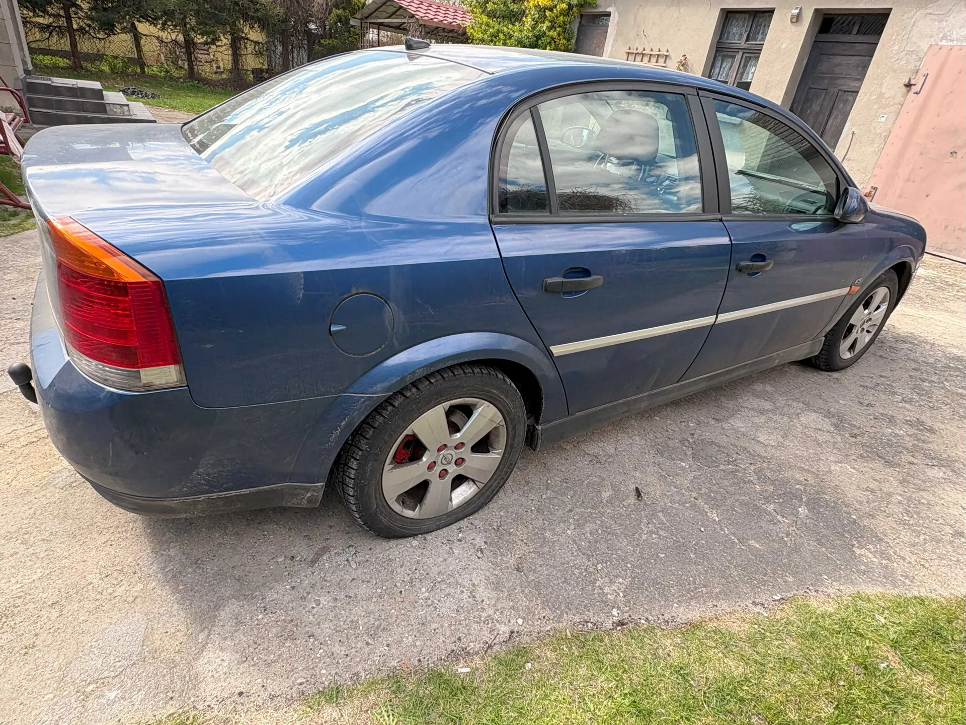 Niebieski samochód osobowy typu sedan zaparkowany na betonowej powierzchni. Widoczna tylna i boczna część pojazdu z czerwonymi światłami tylnymi, srebrnymi felgami i czerwonymi zaciskami hamulcowymi. Samochód jest nieco zabrudzony. W tle dom z brązowymi drzwiami, oknami, zielonymi krzewami i trawnikiem. Na lewo od samochodu schody prowadzące do budynku. Niebo odbija się w szybach pojazdu.