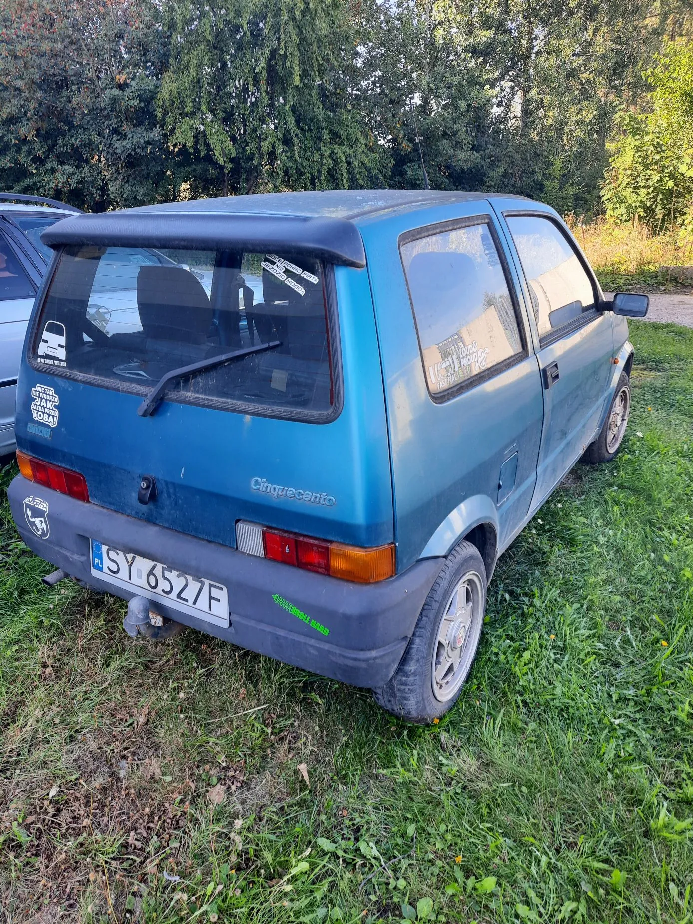 Niebieski, trzydrzwiowy samochód Fiat Cinquecento zaparkowany na trawie. Widoczny jest tył i prawy bok pojazdu. Na klapie bagażnika znajduje się napis 'Cinquecento' oraz polska tablica rejestracyjna o numerze SY 6527F. Na tylnej szybie i karoserii naklejone są różne naklejki. W tle widoczne są zielone drzewa i krzewy.