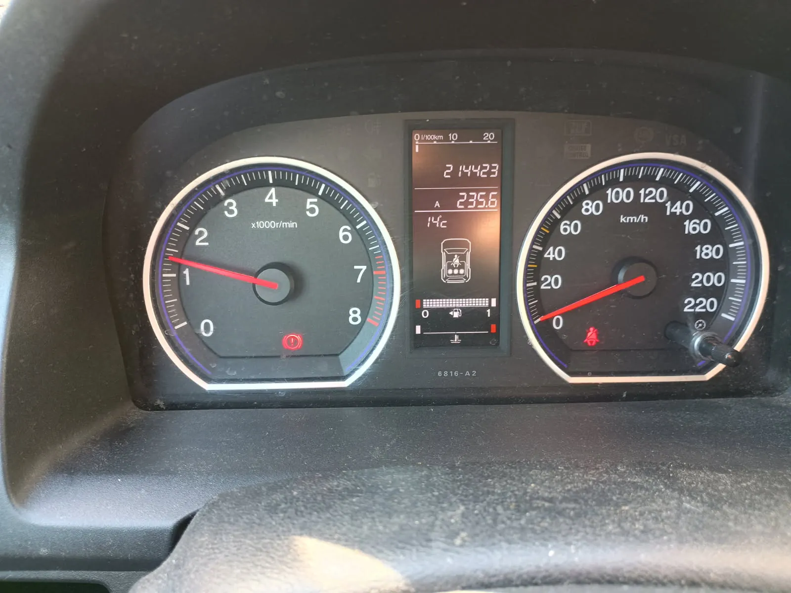 Tablica rozdzielcza samochodu z dwoma głównymi wskaźnikami. Po lewej stronie wskaźnik obrotomieru z zakresem od 0 do 8 (x1000r/min), czerwona igła wskazująca około 1. Po prawej stronie wskaźnik prędkości z zakresem od 0 do 220 km/h, czerwona igła wskazująca 0. W centralnym wyświetlaczu cyfrowym widoczne wartości: 214423 (przebieg), 235.6 (przebieg podróży), 14°C (temperatura zewnętrzna), ikona zbiornika paliwa prawie pustego oraz czerwone światło ostrzegawcze. Na tle czarna deska rozdzielcza z białymi i czerwonymi oznaczeniami.