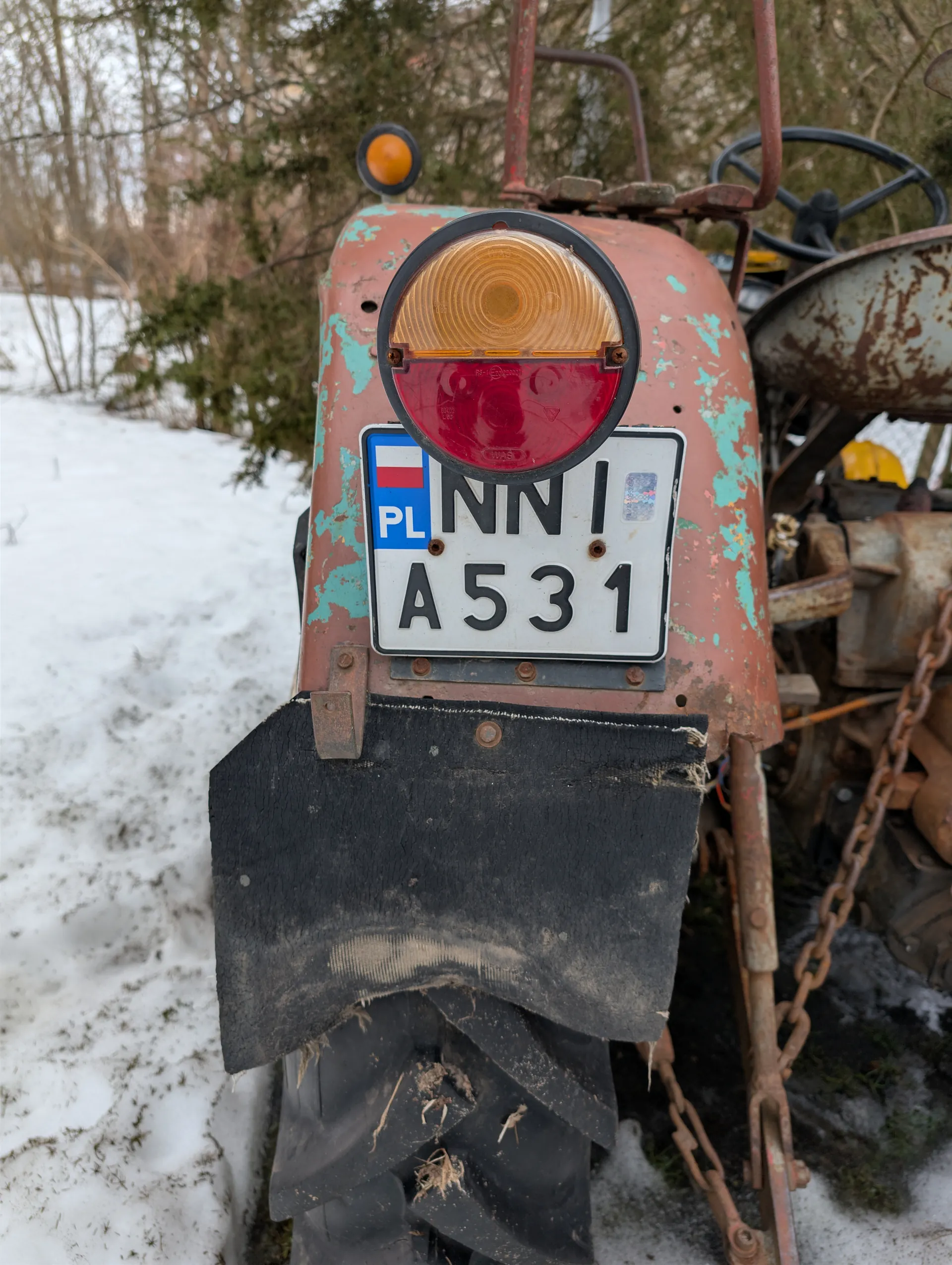 Zdjęcie przedstawia tylną część zardzewiałego traktora z obłupioną pomarańczową farbą i zielonymi plamami. W centrum umieszczona jest biała tablica rejestracyjna z czarnym napisem "PL NN A531", po lewej stronie tablicy widoczny jest niebieski prostokąt z biało-czerwoną flagą Polski. Nad tablicą znajduje się okrągły reflektor z pomarańczową i czerwoną częścią. Poniżej tablicy widoczna jest czarna, zużyta osłona. W tle śnieżna powierzchnia, drzewa bez liści oraz fragmenty innych maszyn rolniczych. Na traktorze widoczne są również rdzewne łańcuchy i kierownica.