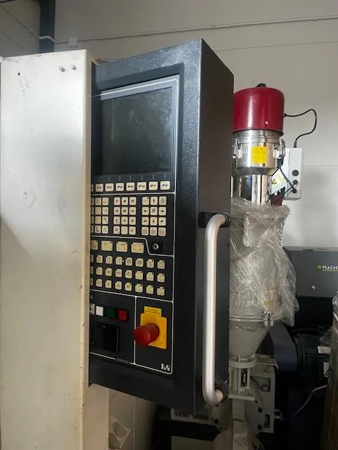 Przemysłowe urządzenie z czarnym panelem sterowania zawierającym ekran, siatkę przycisków z oznaczeniami oraz czerwony przycisk awaryjny na żółtym tle. Po prawej stronie panelu widoczny czerwony cylinder z żółtą etykietą, przewodami i częścią owiniętą przezroczystą folią. W tle biała ściana i fragment innej maszyny z częściowo widocznym napisem 'MACH'.