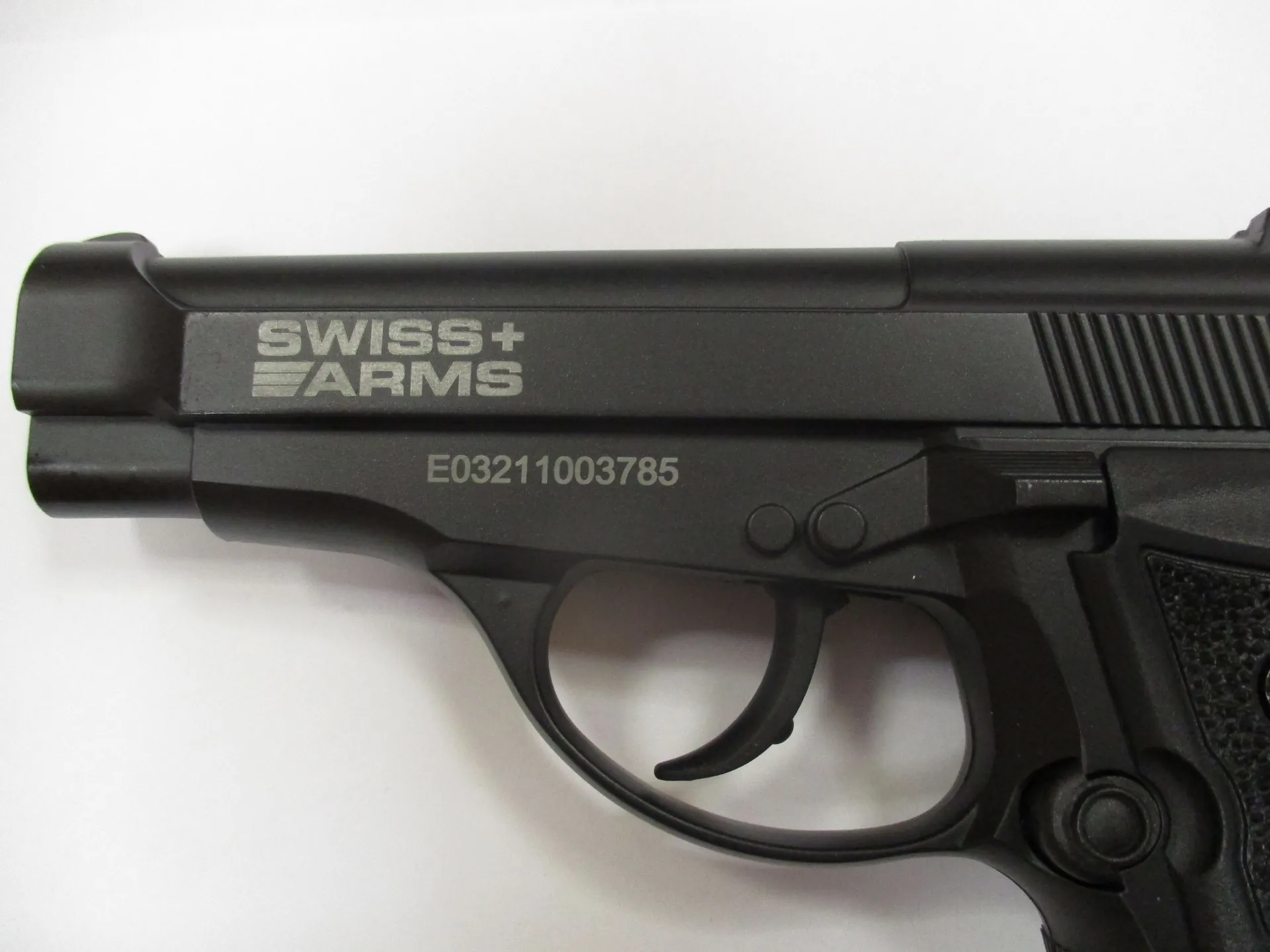 Zbliżenie na czarną broń palną typu pistolet. Na zamku widnieje napis 'SWISS+ ARMS', a poniżej, na ramie, numer seryjny 'E03211003785'. Widoczny jest spust oraz część rękojeści z chropowatą fakturą.