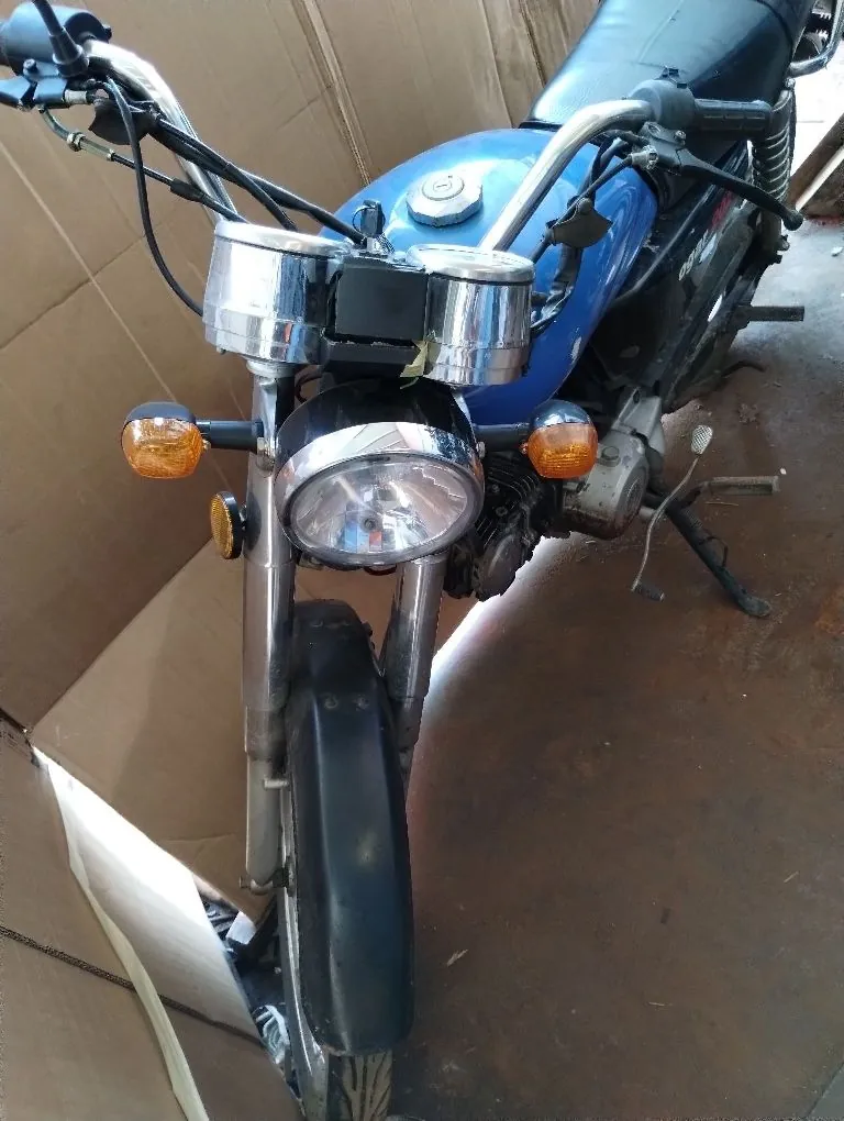 Widok z przodu na niebieski motocykl zaparkowany w pomieszczeniu na brązowej podłodze. Widoczny jest przedni reflektor, dwa pomarańczowe kierunkowskazy, chromowane zegary na kierownicy oraz przedni błotnik w kolorze czarnym.