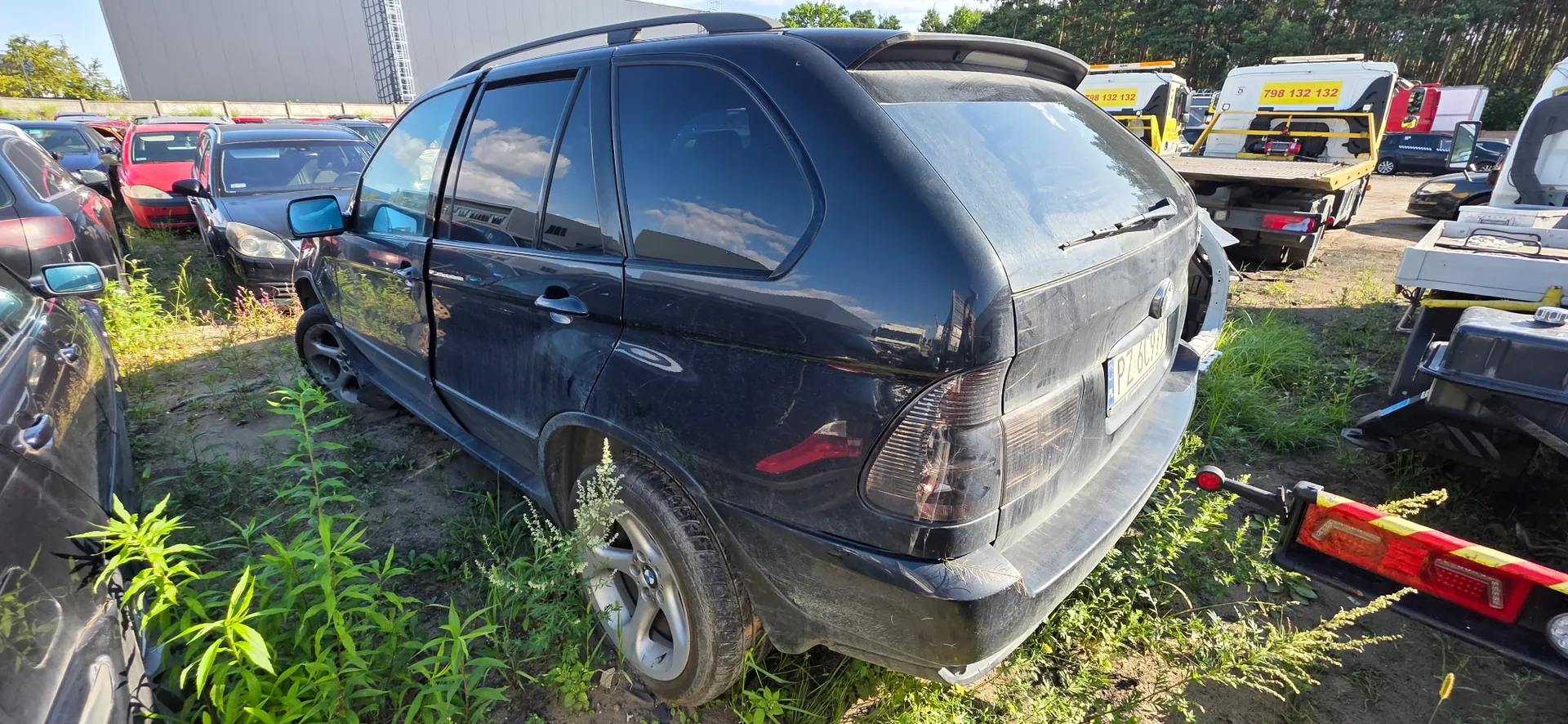 Czarny samochód typu SUV zaparkowany na trawiastym placu. Pojazd jest zakurzony i uszkodzony z tyłu, widoczna jest tablica rejestracyjna PZ 869... W tle znajdują się inne samochody oraz białe ciężarówki z żółtymi napisami, w tym numerem telefonu 798 132 132.