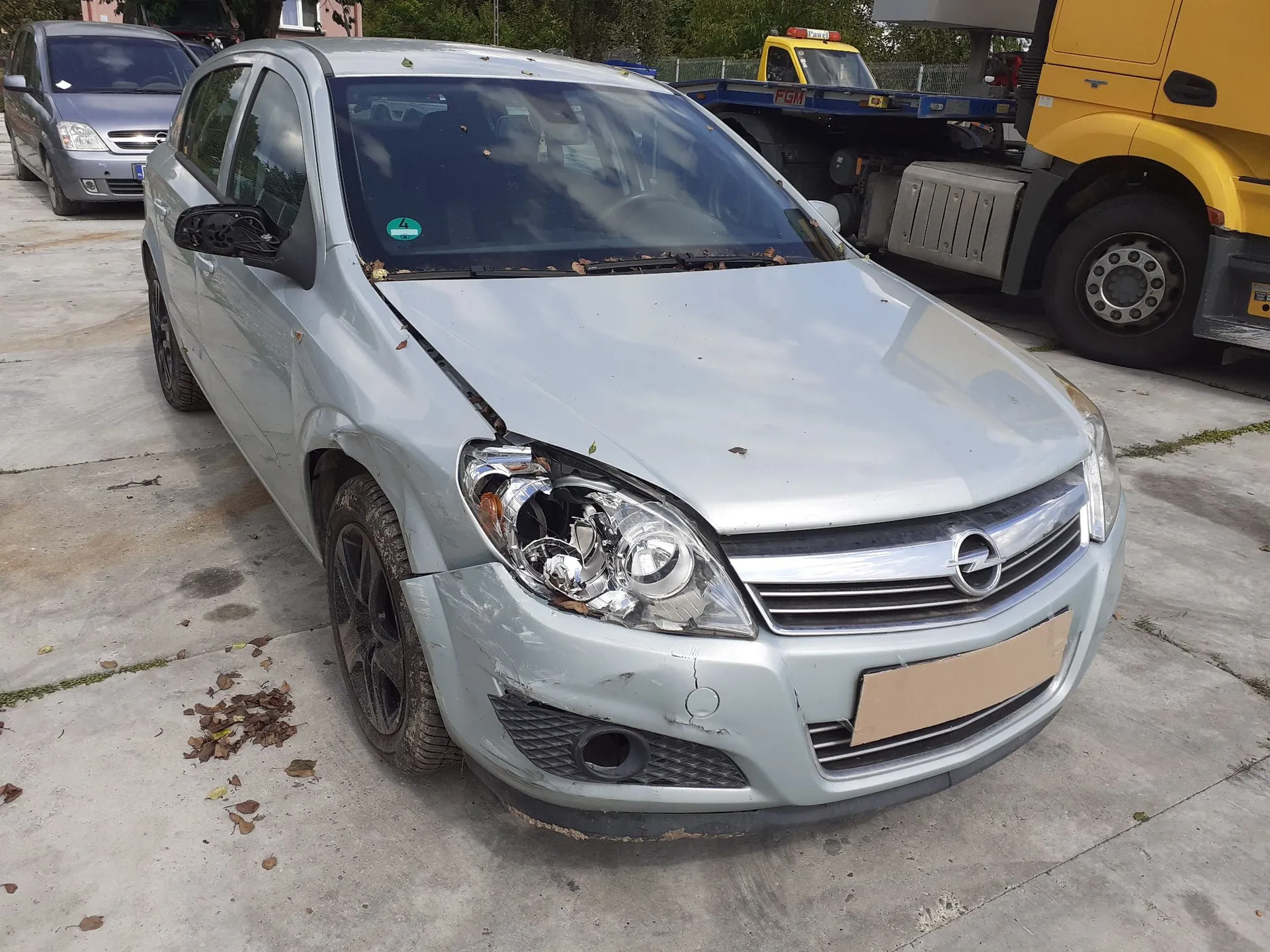 Srebrny samochód osobowy marki Opel z widocznymi uszkodzeniami przodu po prawej stronie. Rozbita jest obudowa przedniego reflektora, a zderzak oraz błotnik są wgniecione. Auto stoi na betonowym placu obok żółtej ciężarówki i szarego vana.