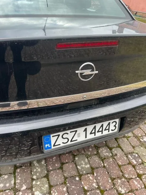 Tylne widok czarnego samochodu z okrągłym logo z piorunem w centrum, czerwonym światłem tylnym oraz białą tablicą rejestracyjną z niebieskim pasem zawierającym napis "PL" i czarnymi literami i cyframi "ZSZ 14443". Samochód stoi na brukowanej powierzchni. W tle widoczne są odbicia osób i budynków w szybie.