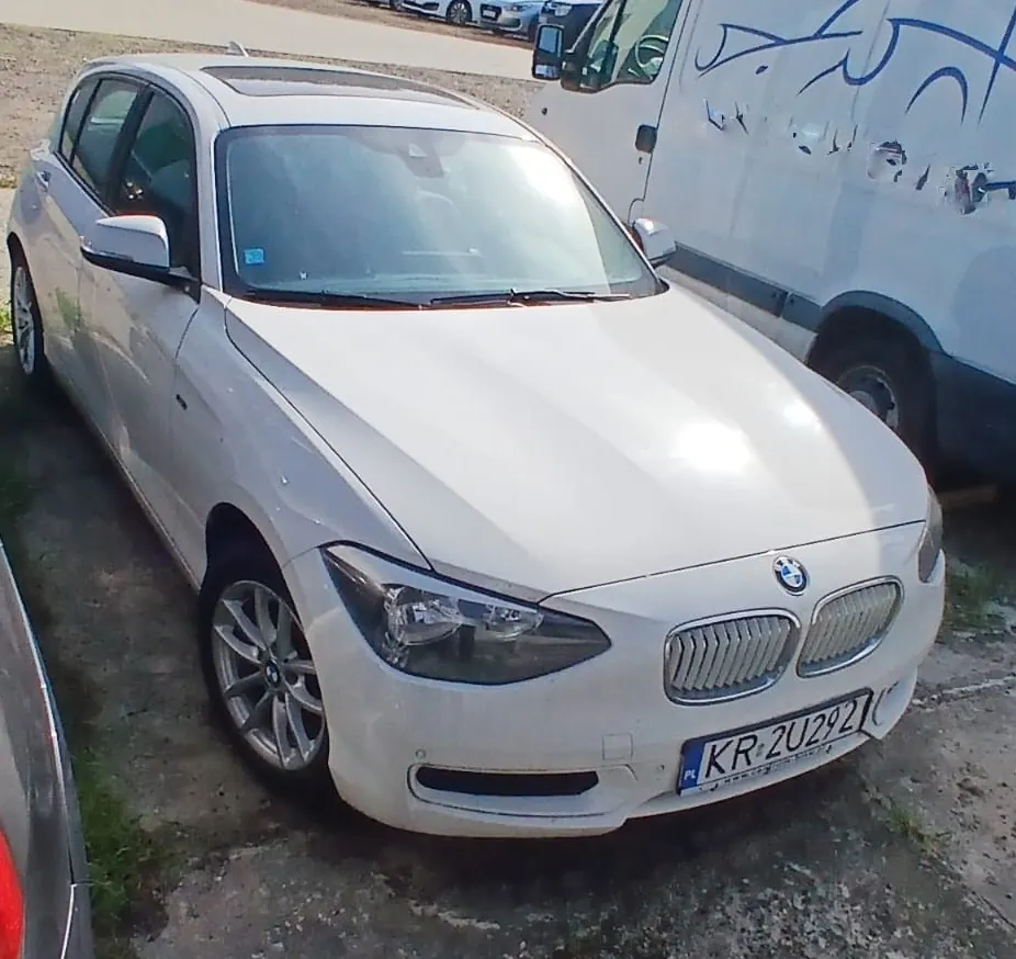 Biały samochód osobowy marki BMW zaparkowany na utwardzonym placu. Pojazd jest widoczny z przodu i z boku. Na masce widoczne jest logo BMW, a z przodu znajduje się tablica rejestracyjna o numerze KR 2U292. Obok samochodu zaparkowany jest biały furgon z niebieskimi napisami na boku.