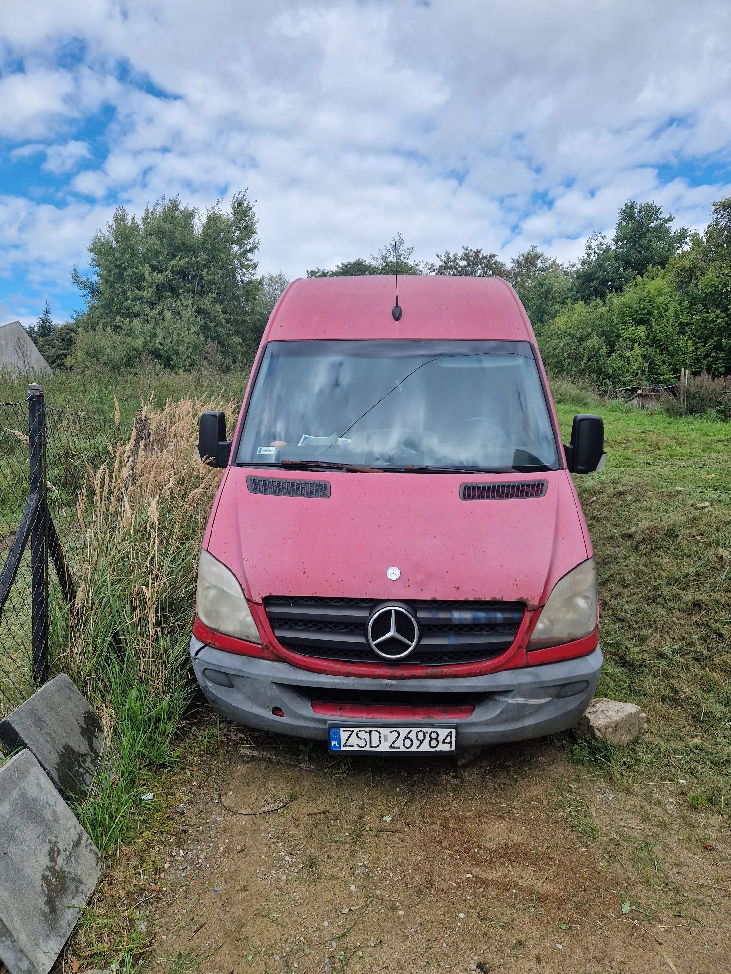 Czerwony furgon marki Mercedes-Benz zaparkowany na piaszczystej drodze w otoczeniu zieleni i wysokich traw. Pojazd ma polską tablicę rejestracyjną o numerze ZSD 26984. W tle widoczne są drzewa oraz niebo z białymi chmurami.