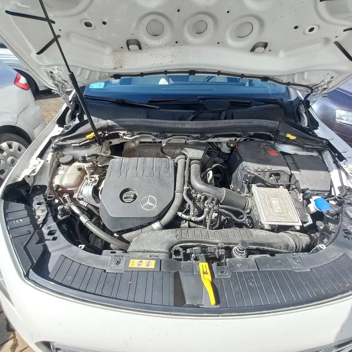 Widok z góry na otwartą maskę białego samochodu, ukazujący komorę silnika. W centrum znajduje się czarna plastikowa osłona silnika z logo marki Mercedes-Benz. Widoczne są elementy mechaniczne, przewody, akumulator po prawej stronie oraz zbiornik płynu chłodniczego po lewej. Na pierwszym planie znajduje się czarny plastikowy panel z żółtym uchwytem bagnetu do pomiaru poziomu oleju.