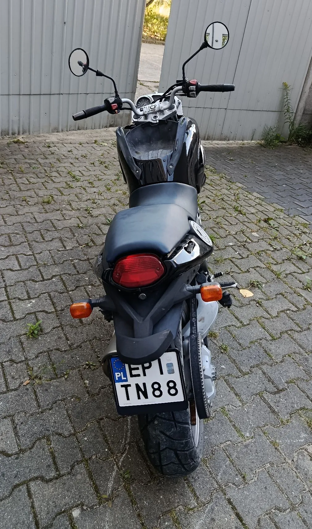 Widok z tyłu na czarny motocykl zaparkowany na kostce brukowej przed szarym, metalowym ogrodzeniem. Motocykl posiada czerwoną tylną lampę, pomarańczowe kierunkowskazy oraz polską tablicę rejestracyjną o numerze EP1 TN88.