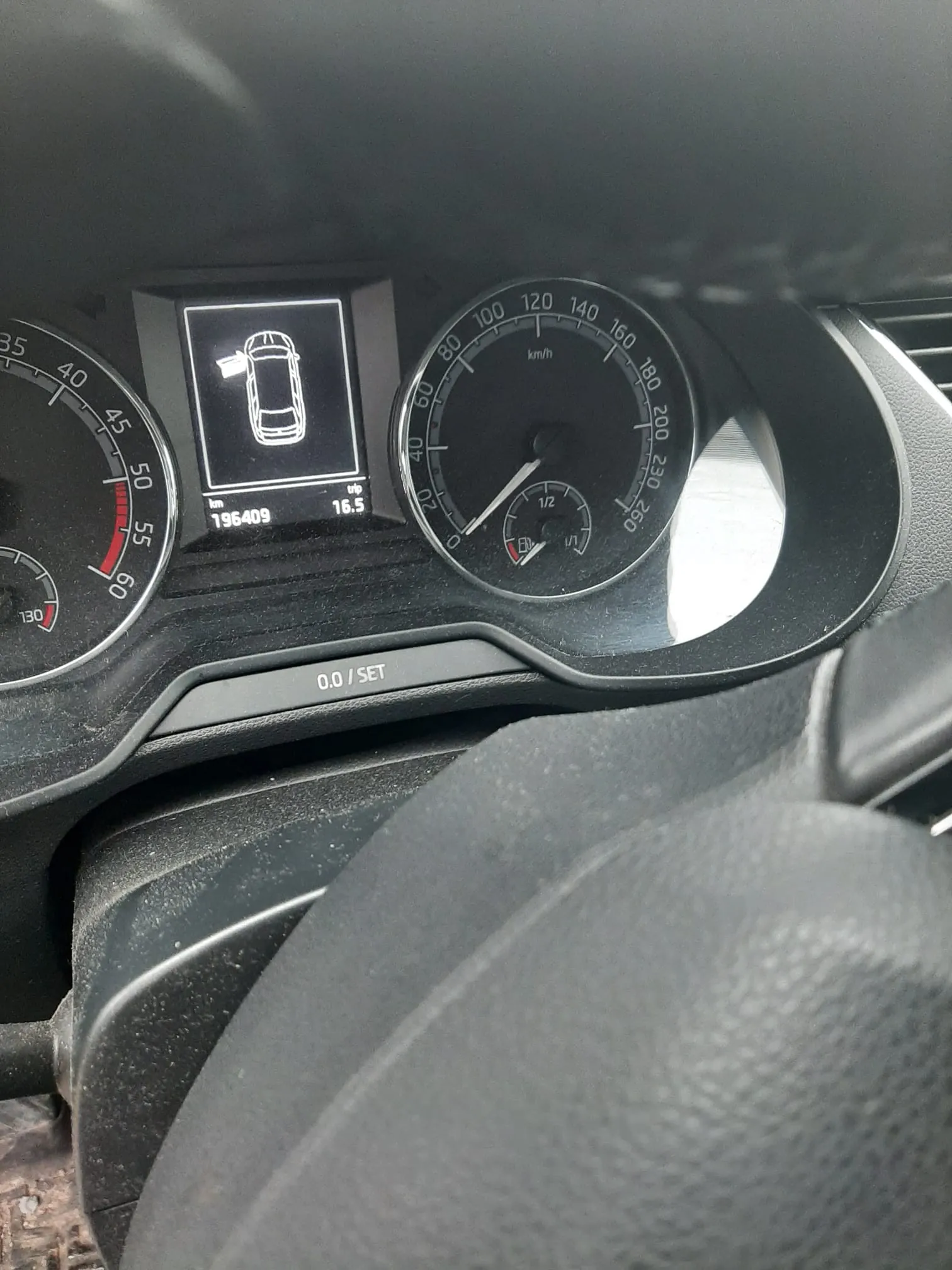 Zbliżenie na deski rozdzielcze samochodu. Po lewej stronie widoczny wskaźnik prędkości z czerwonym segmentem, po prawej główny wskaźnik prędkości w km/h (0-260) z podwskaźnikiem paliwa. W centrum wyświetlacz cyfrowy z ikoną samochodu z otwartą klapą bagażnika, przebiegiem 196409 km i licznikiem podróży 16.5. Poniżej wyświetlacz napis 0.0 / SET. W dolnej części widoczna część kierownicy.