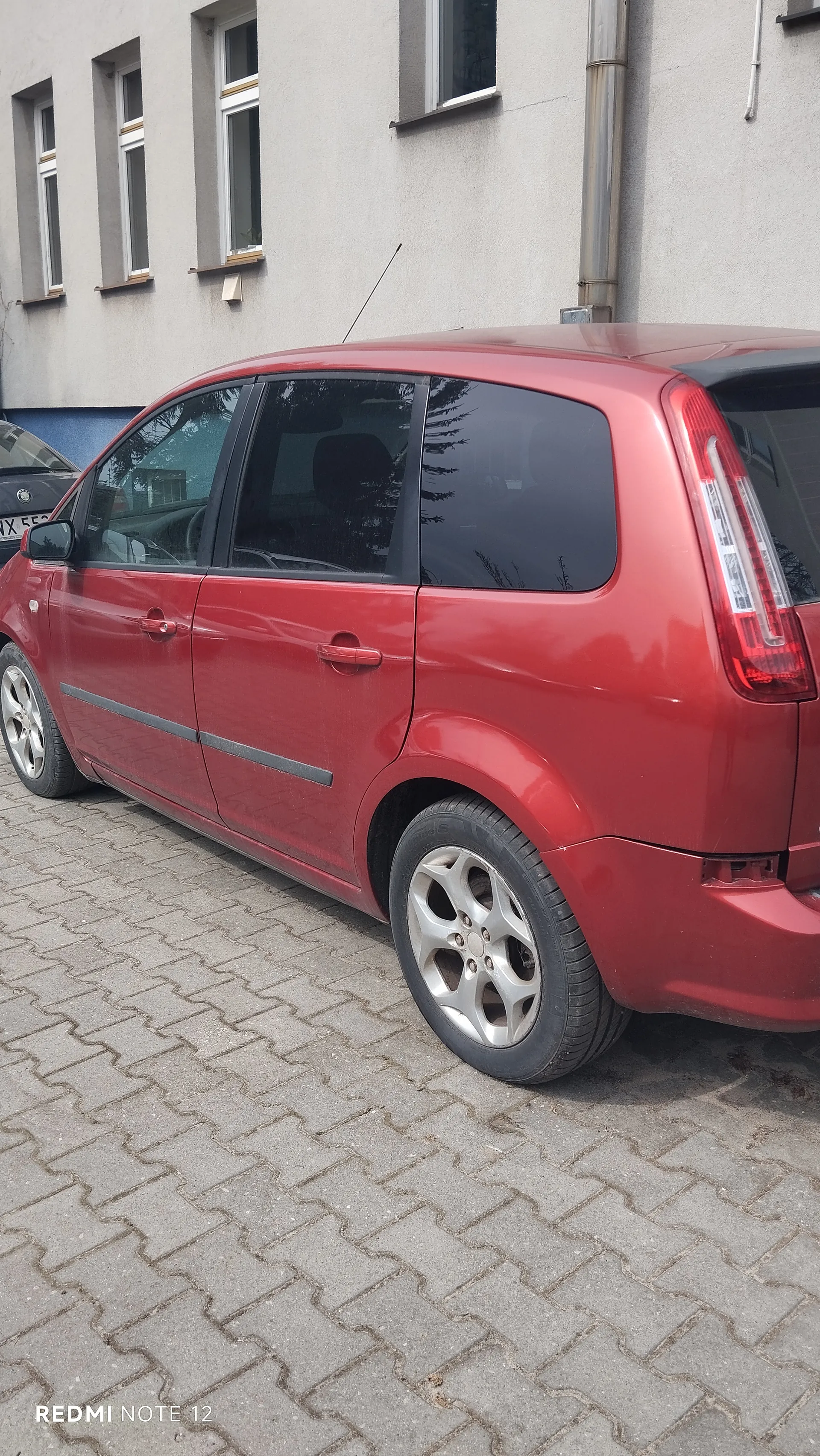 Czerwony samochód typu hatchback zaparkowany na brukowanej powierzchni przed budynkiem o szarej elewacji z czterema oknami. Widoczne są czarne szyby, srebrne felgi, tylny reflektor i antena na dachu. W tle częściowo widoczny inny samochód z tablicą rejestracyjną zaczynającą się na X 55. W lewym dolnym rogu napis REDMI NOTE 12.