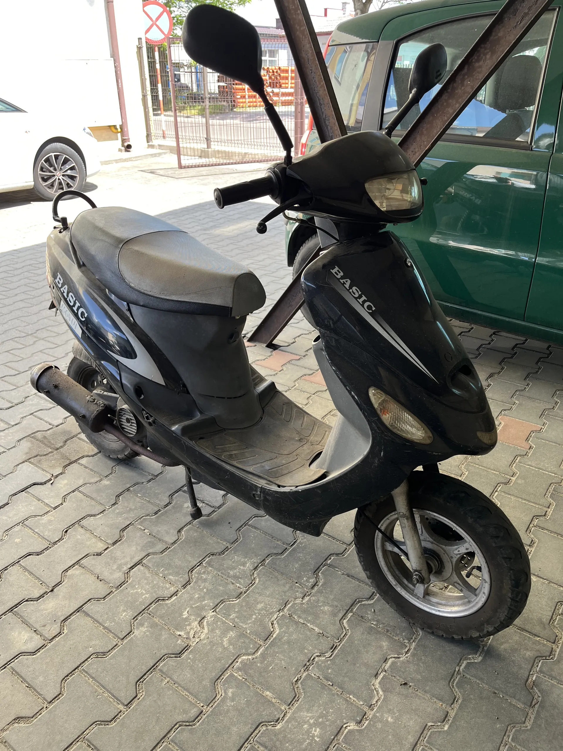 Czarny skuter z napisem BASIC na nadwoziu zaparkowany na chodniku z geometrycznym wzorem. Obok skutera widoczny zielony samochód. W tle biały samochód, znak zakazu wjazdu oraz budynki. Skuter posiada lusterka, reflektory i widoczne koła.