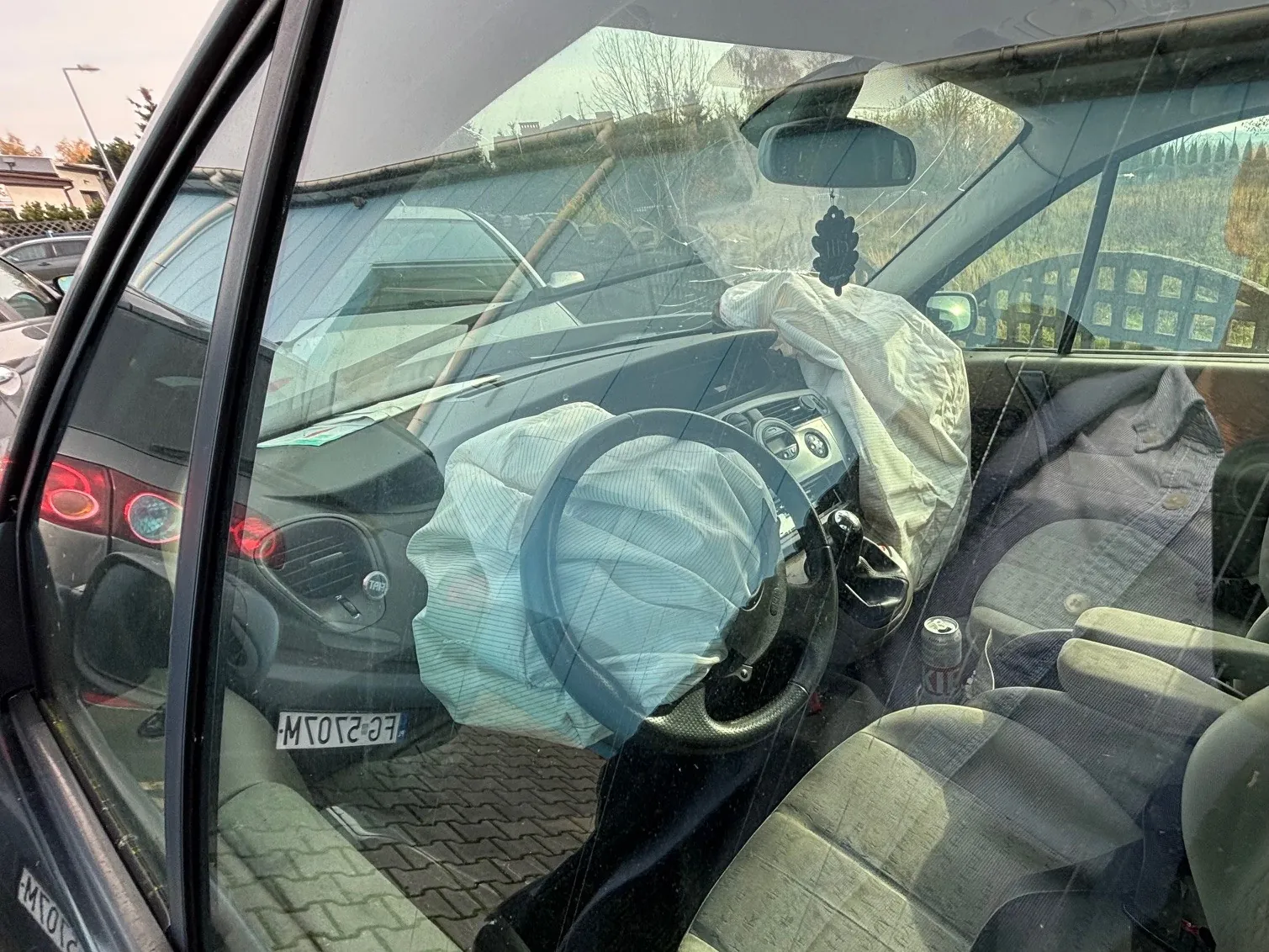 Wnętrze samochodu z rozwiniętymi poduszkami powietrznymi na kierownicy i pasażerskim siedzeniu. Widoczna tablica rejestracyjna FE-2107M, szare siedzenia, kierownica, deska rozdzielcza oraz lusterko wsteczne z wiszącym aromatyzatorem. Poza samochodem widoczne są inne pojazdy, budynki i drzewa. Na siedzeniu pasażera stoi puszka.