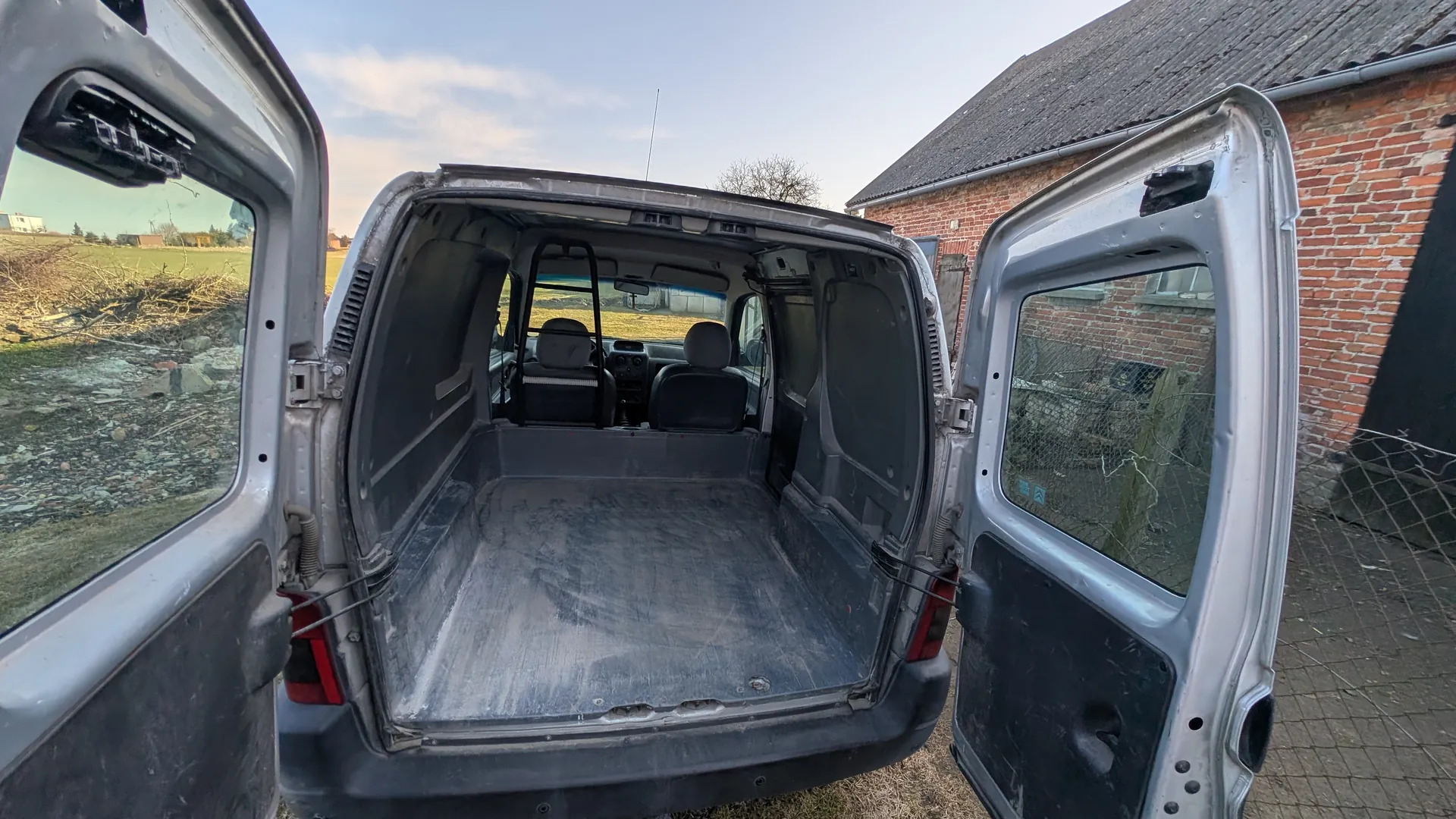 Widok na tył otwartego furgonu z pustym przestrzenią ładunkową. Drzwi tylne są rozchylone, widać czarne siedzenia kierowcy i pasażera oraz deski rozdzielczą. Po prawej stronie ceglany budynek z dachem krytym dachówką, po lewej pole z suchą roślinnością i gruzem. Niebo częściowo zachmurzone. Na szybie prawego drzwi widoczny mały niebieski naklejony znak.
