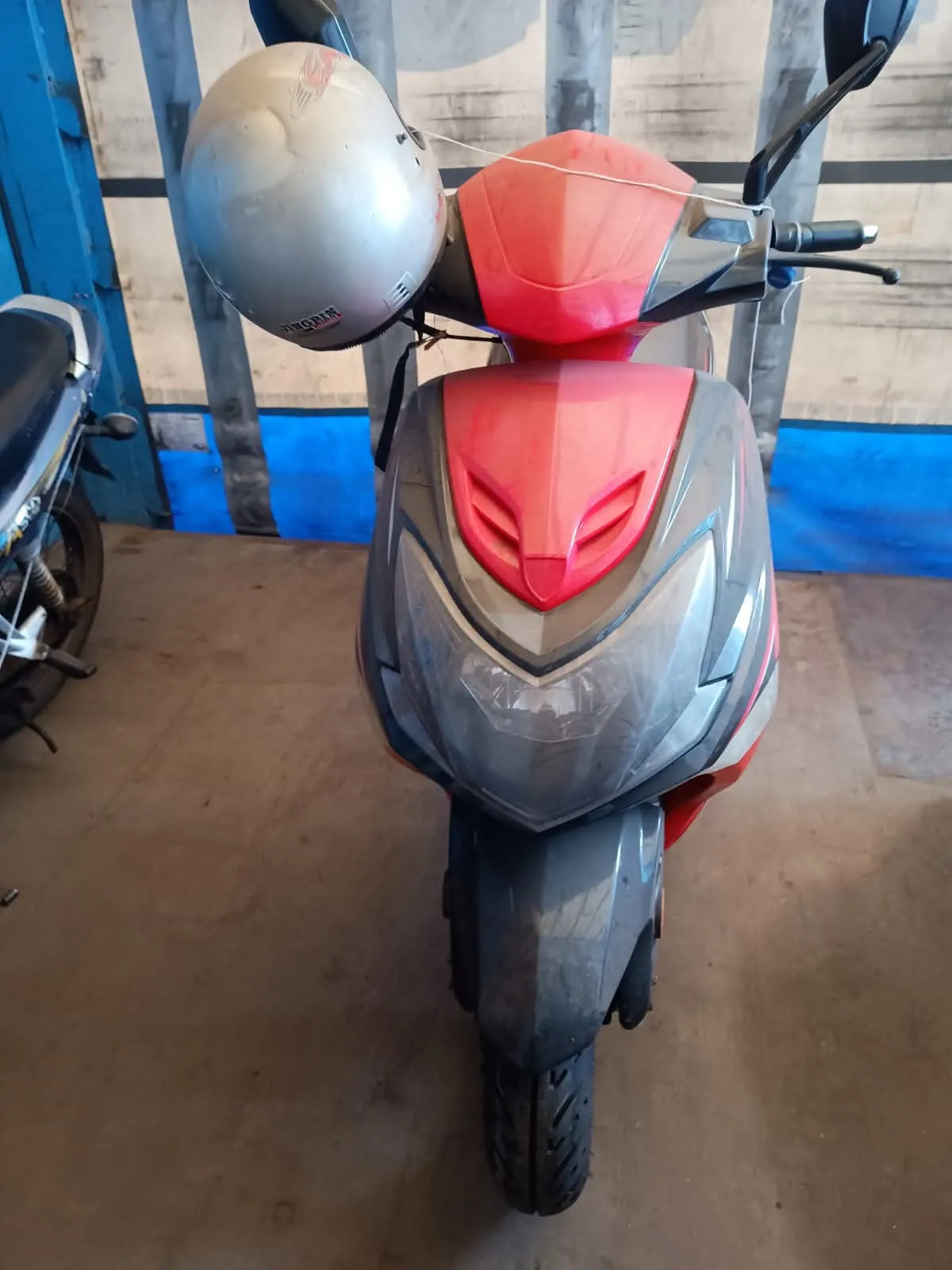 Zdjęcie przedstawiające przód czerwono-czarnego skutera zaparkowanego w garażu lub magazynie. Na lewym lusterku skutera zawieszony jest srebrny kask motocyklowy. W tle widoczna jest niebieska ściana i fragment innego motocykla po lewej stronie.