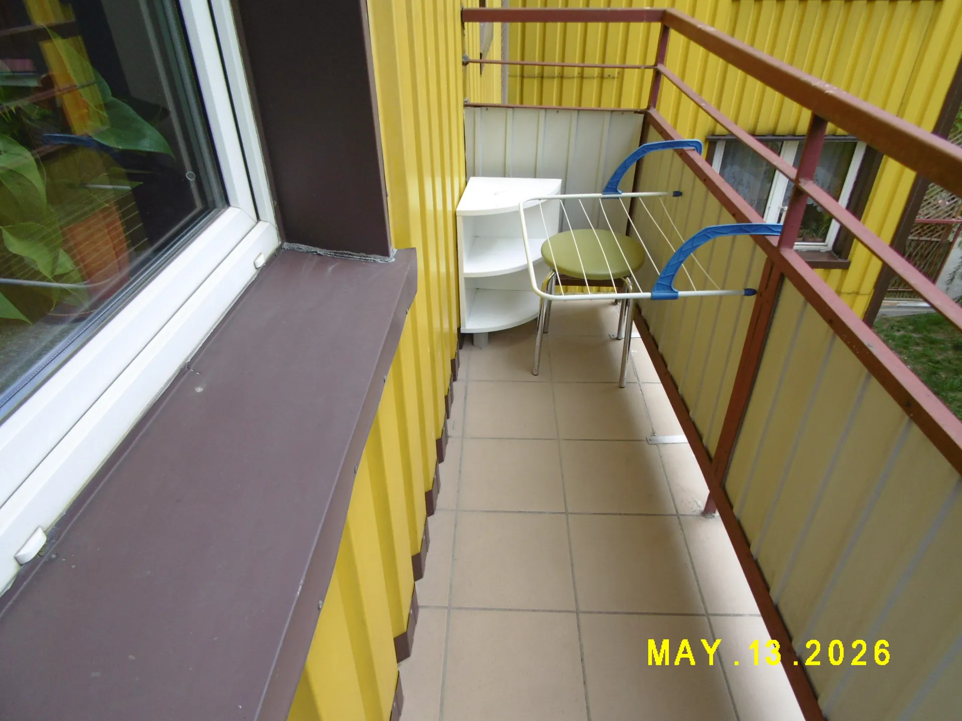Widok na wąski balkon z żółtymi ścianami i brązową balustradą. Na balkonie stoi biały regał z półkami oraz rozkładana suszarka na pranie z niebieskimi elementami, na której leży zielony przedmiot. Podłoga wyłożona jest beżowymi płytkami. W lewym dolnym rogu znajduje się brązowy parapet okienny. W prawym dolnym rogu widnieje żółty napis daty: MAY 13 2026.
