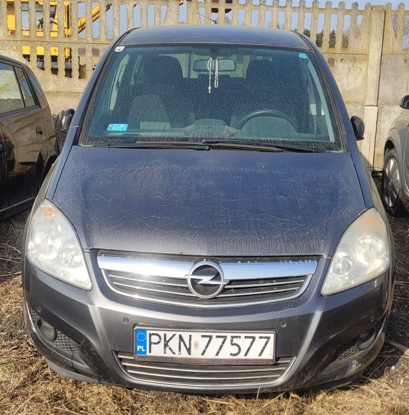 Przód ciemnoszarego samochodu marki Opel z widocznym logo na masce. Na masce widoczne są pęknięcia i zabrudzenia. Tablica rejestracyjna z białym tłem, niebieskim pasem z napisem 'PL' po lewej stronie oraz czarnym tekstem 'PKN 77577'. Po obu stronach samochodu częściowo widoczne są inne pojazdy. Tło stanowi betonowy mur z drewnianym ogrodzeniem na górze. Podłoga pokryta suchą trawą.