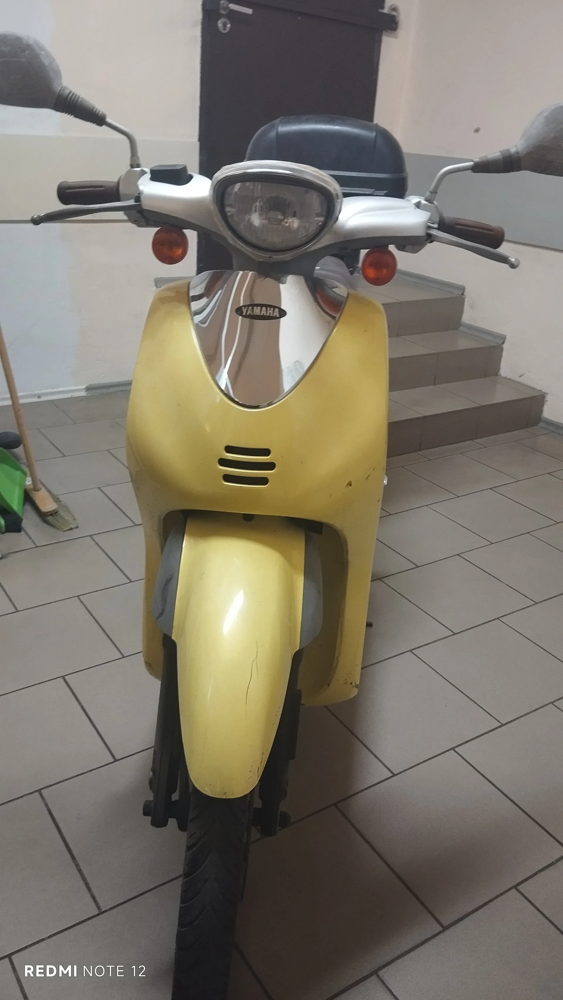 Zdjęcie przedstawia żółty skuter marki Yamaha z przodu. Skuter ma srebrną maskę z logo Yamaha, okrągły reflektor, dwa pomarańczowe kierunkowskazy oraz czarne lusterka boczne z brązowymi uchwytami. W tle widoczne są schody z płytek, ciemne drzwi oraz miotła z zielonym zbiornikiem po lewej stronie. Na dole zdjęcia znajduje się napis REDMI NOTE 12. Podłoga wyłożona jest szarymi płytkami.