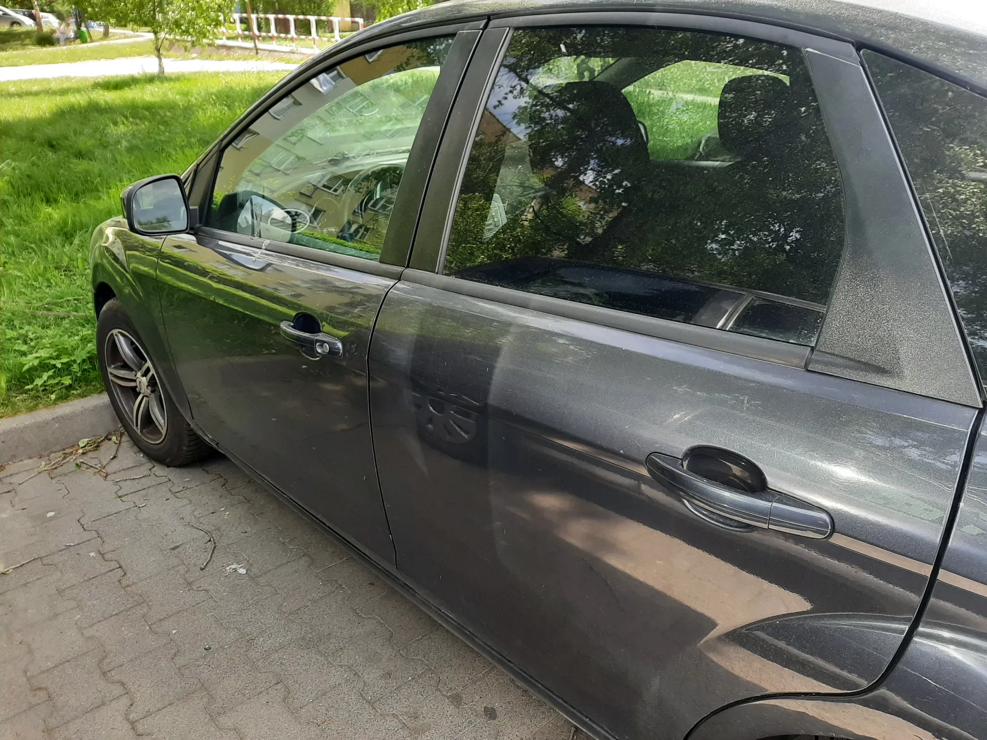Czarny samochód osobowy zaparkowany na brukowanej powierzchni. Widoczne są boczne drzwi, lusterko boczne oraz odbicia w szybach: budynki, drzewa i zielona trawa. W tle trawa, drzewo i fragment drogi z białoczerwonymi barierami.