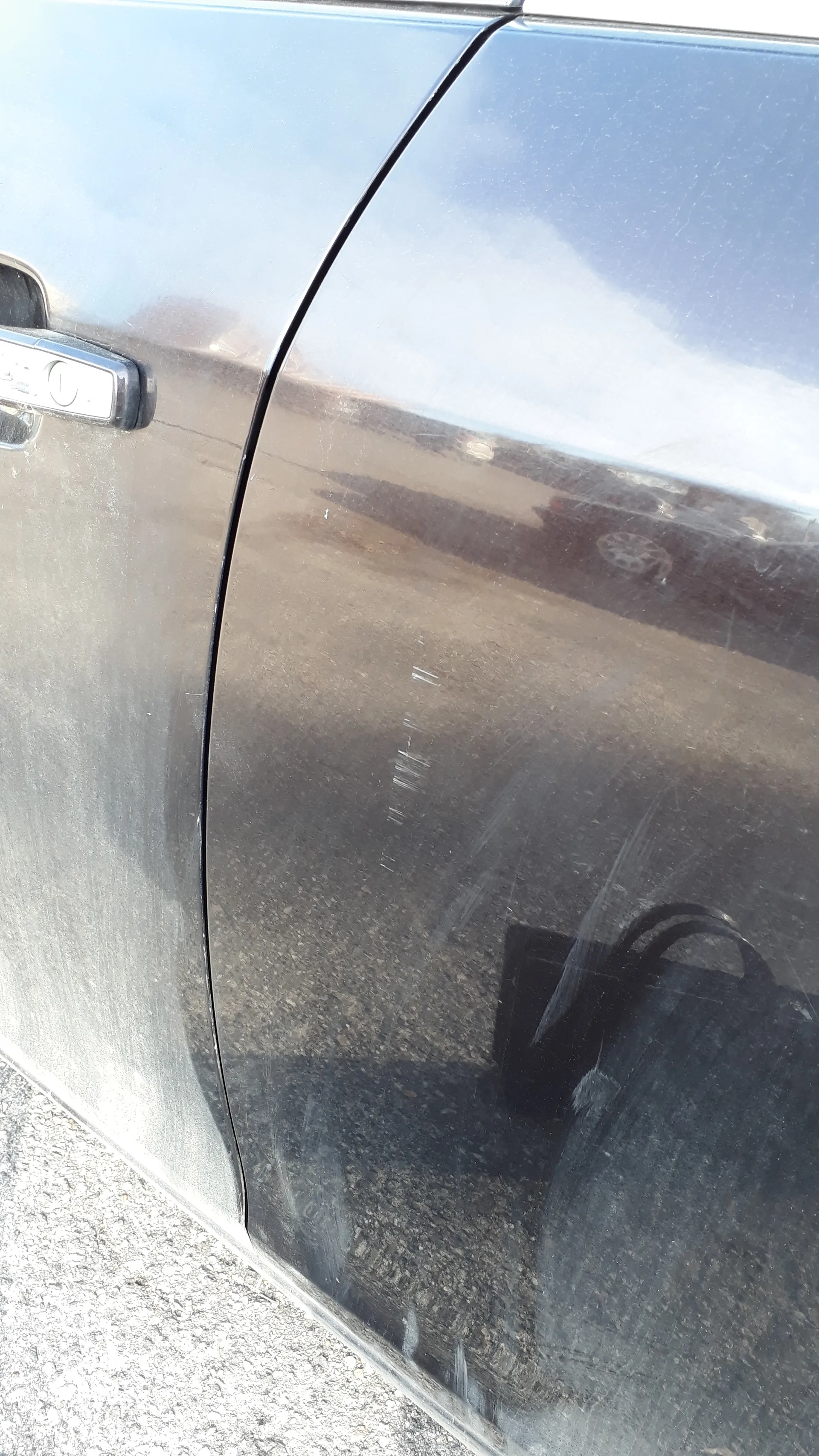 Zbliżenie na ciemnoszary bok samochodu z metalową klamką drzwi. Na powierzchni samochodu widoczne są odbicia: drogi, żwiru, oddalonego pojazdu oraz częściowo zachmurzonego nieba.