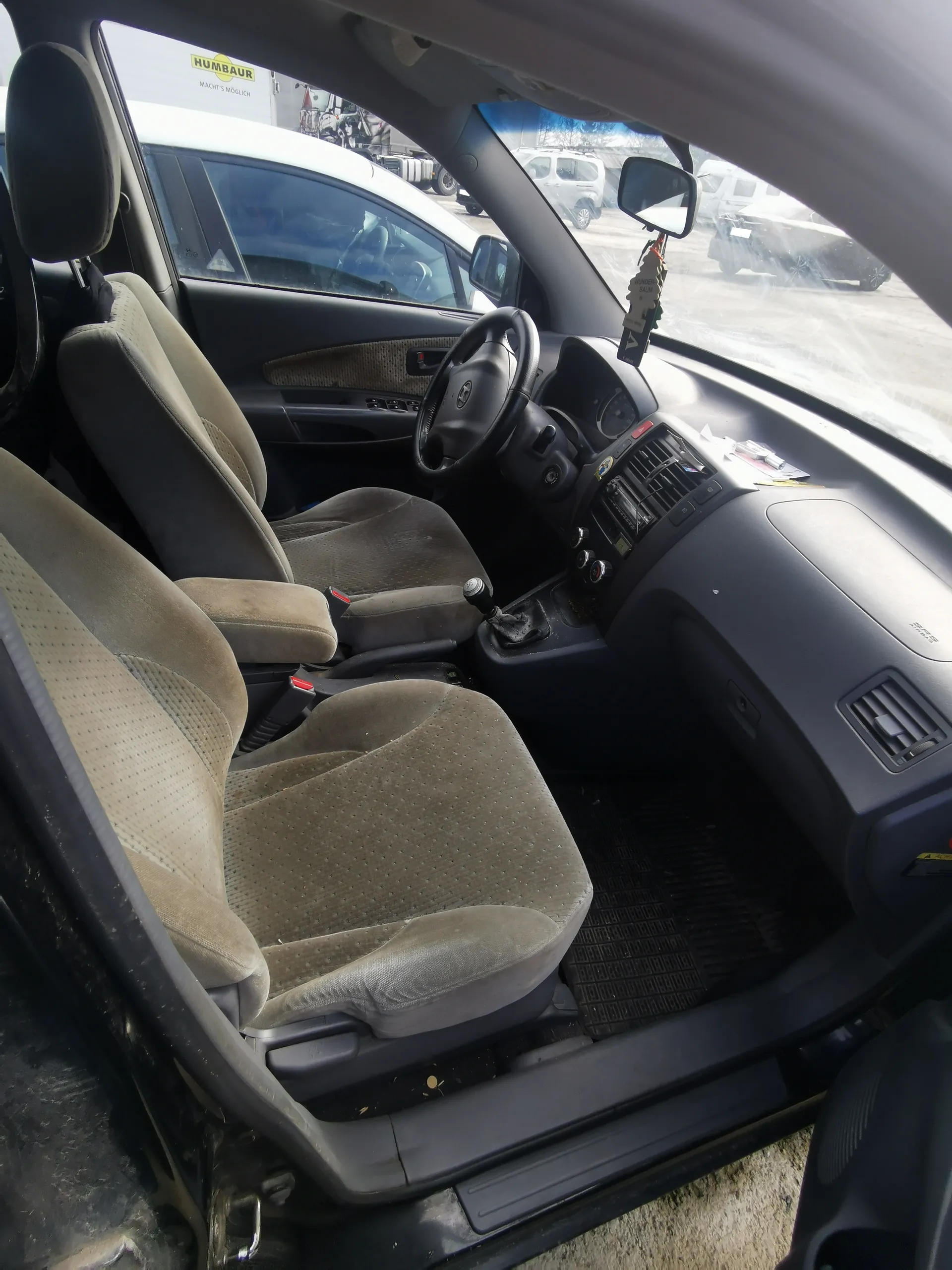 Wnętrze samochodu z beżowymi tkaninowymi fotelami, kierownicą po lewej stronie, dźwignią zmiany biegów i czarną deską rozdzielczą. Drzwi kierowcy są otwarte. Na zewnątrz widoczne są inne pojazdy oraz tablica z napisem "HUMBAUR" i podpisem "MACHT'S MÖGLICH". W lusterku wstecznym wiszą przedmioty. Podłoga pokryta czarnymi matami.