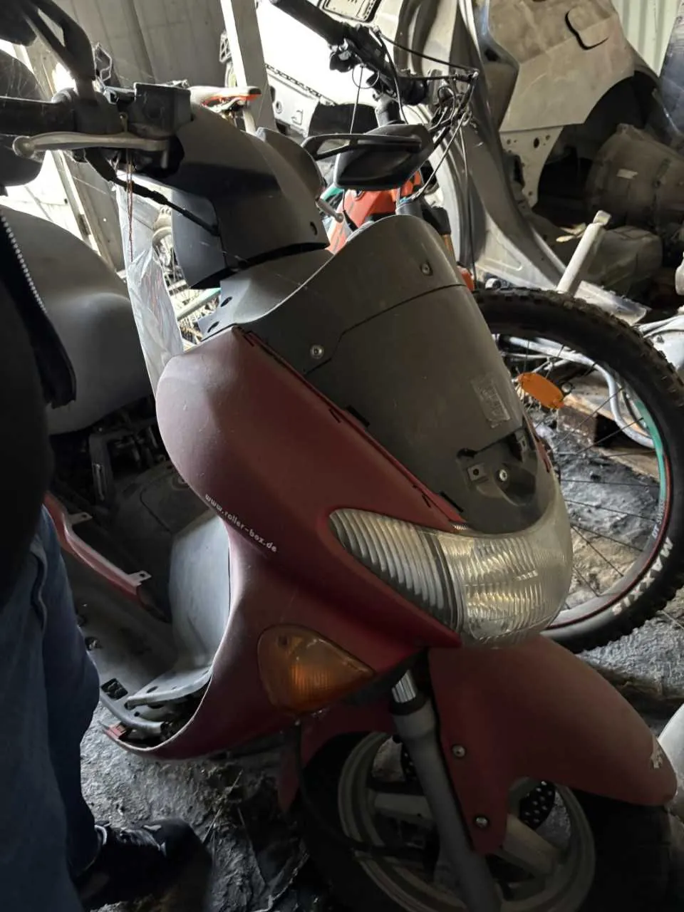 Czerwono-szary skuter z przodu, z widocznym reflektorem, kierunkowskazem i napisem www.roller-box.de na bocznej części. W tle fragmenty innych pojazdów, w tym koło roweru z odblaskiem oraz części motocykla. Po lewej stronie widoczna część nogi osoby w niebieskich dżinsach i czarnym buciku. Podłoga z betonu, otoczenie przypomina warsztat lub magazyn.