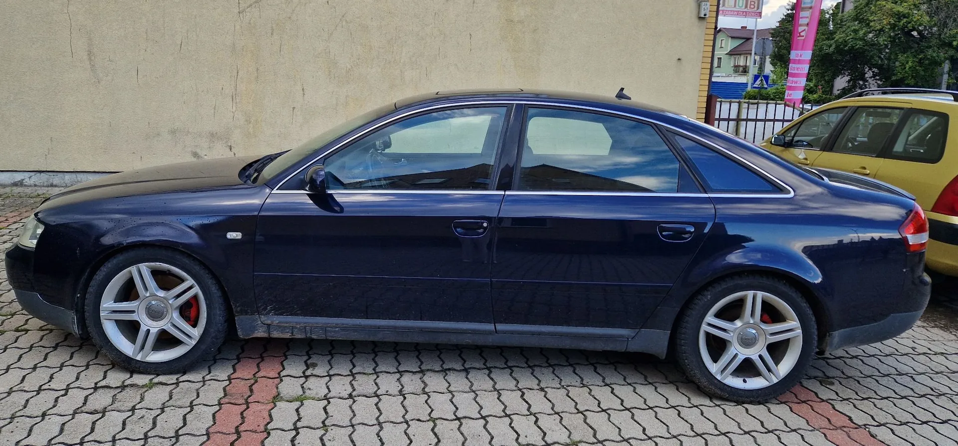 Profil ciemnoniebieskiego samochodu osobowego typu sedan zaparkowanego na kostce brukowej. Samochód ma srebrne felgi z pięcioma szerokimi ramionami i czerwone zaciski hamulcowe. W tle widoczna jest jasna ściana budynku oraz fragment żółtego samochodu po prawej stronie.