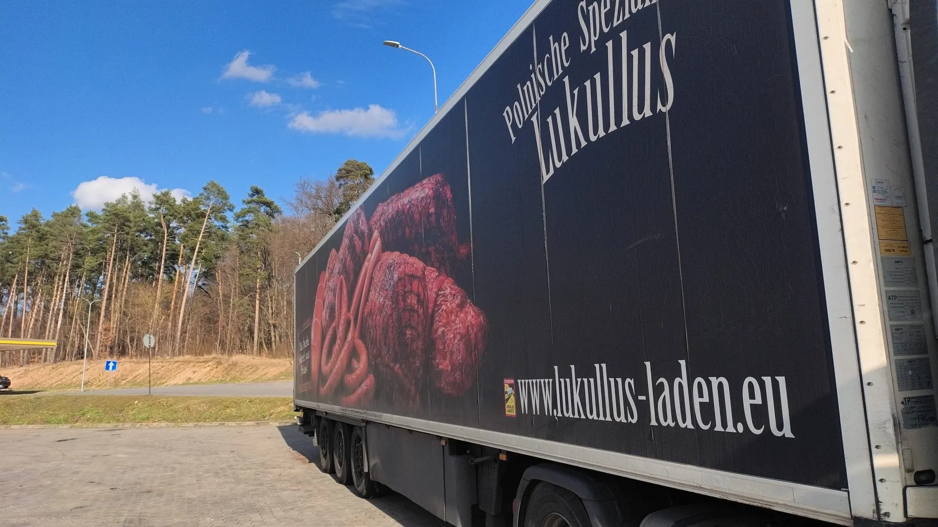 Czarny naczepa ciężarówki z reklamą przedstawiającą mięsne produkty, w tym kiełbasy i suszone mięso. Na naczepie widoczny biały tekst: 'Polnische Spezialitäten Lukullus' oraz adres strony internetowej 'www.lukullus-laden.eu'. W tle niebo z kilkoma chmurami, las iglasty, droga, latarnia uliczna i znaki drogowe. Naczepa stoi na brukowanej powierzchni.