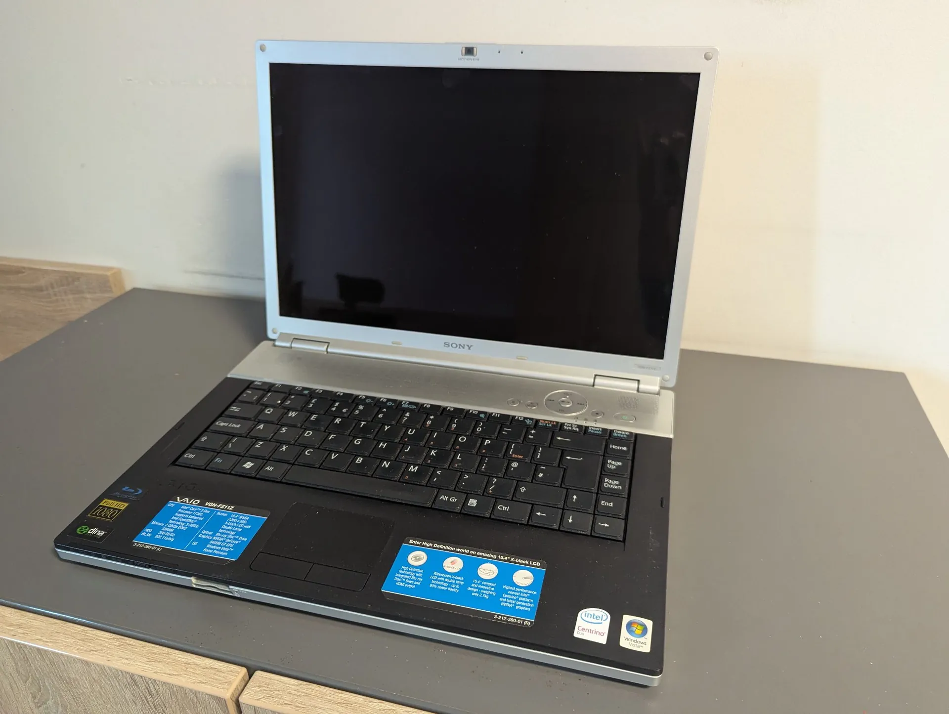 Srebrno-czarny laptop marki Sony VAIO otwarty i ustawiony na szarym blacie. Urządzenie ma srebrną ramkę ekranu i czarną obudowę klawiatury. Na obudowie poniżej klawiatury znajdują się naklejki informacyjne: niebieska z opisem specyfikacji, żółta z napisem 'VAIO', zielona z logo 'dina' oraz naklejki 'Intel Centrino' i 'Windows Vista'. Ekran laptopa jest wyłączony.