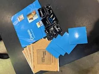 Na ciemnym stole leżą różne przedmioty: niebieska teczka z napisem 'proteQ' i małymi zdjęciami, czarne opakowania z nieczytelnym tekstem, niebieskie karty oraz brązowe koperty z etykietą.