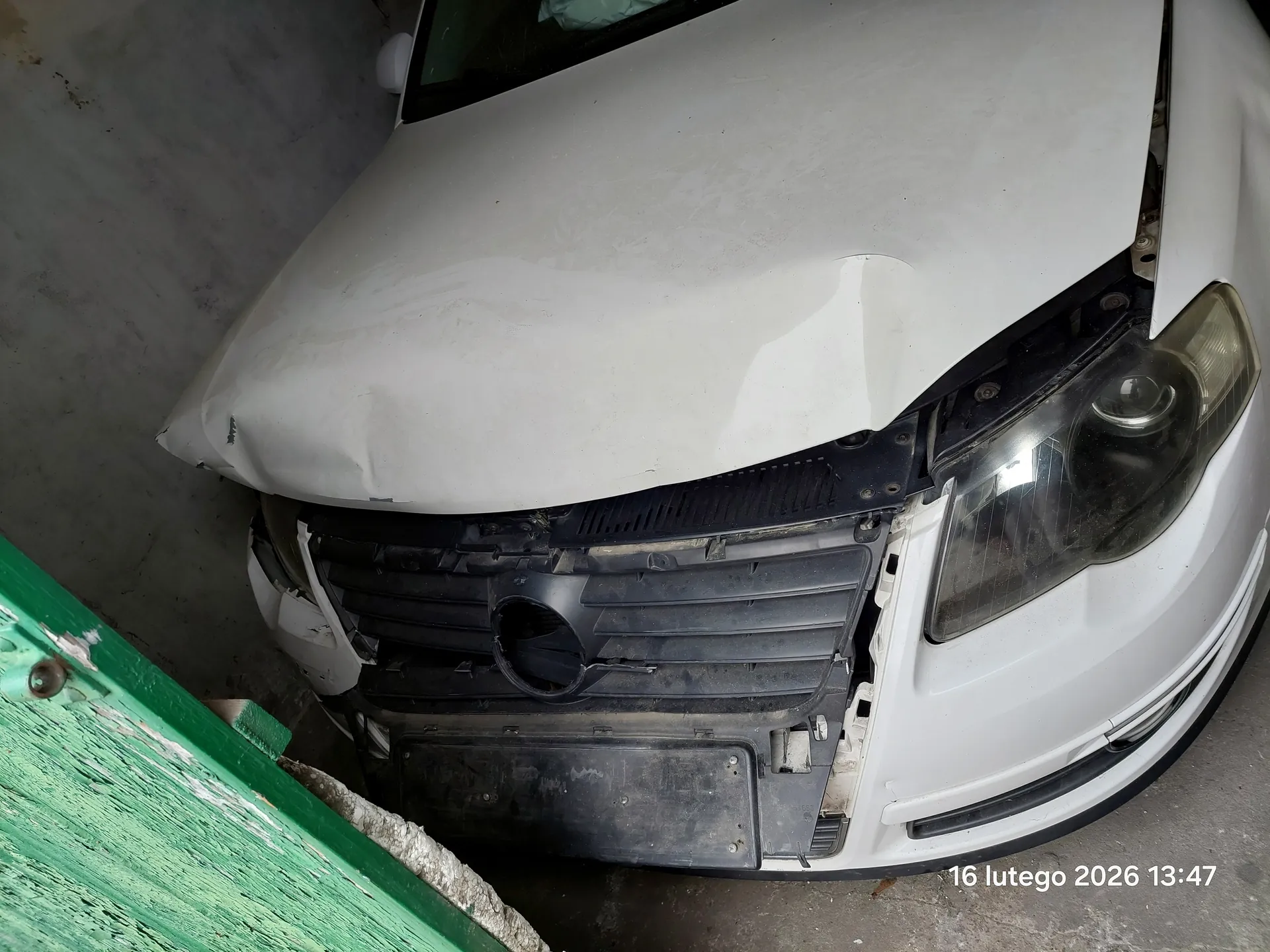 Zdjęcie przedstawia uszkodzony biały samochód z przodu. Widoczna jest zgnieciona maska, uszkodzona atrapa chłodnicy oraz prawe reflektory. Po lewej stronie obrazu znajduje się zielona, zarysowana konstrukcja. Tło stanowi betonowa ściana. W prawym dolnym rogu widoczny jest tekst: 16 lutego 2026 13:47.