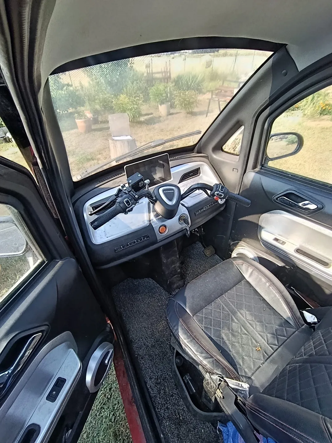 Wnętrze małego pojazdu elektrycznego lub mikrosamochodu. Widoczny jest czarny fotel z rombowym przeszyciem, deska rozdzielcza z kierownicą przypominającą tę z motocykla, na której zamontowany jest ekran tabletu. Przez przednią szybę widać słoneczny ogród z roślinami w doniczkach i trawnikiem.