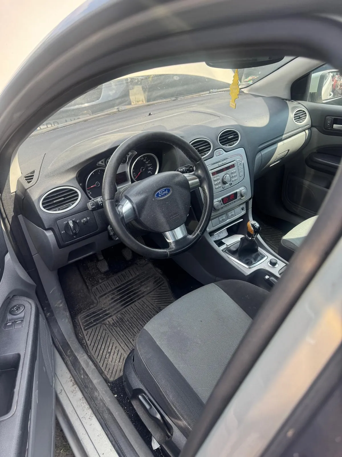 Widok z wnętrza samochodu marki Ford, ukazujący deskę rozdzielczą, kierownicę z logo Ford, fotele oraz lewarek zmiany biegów. Wnętrze jest w kolorach szarych i czarnych. Widoczny jest panel radia oraz wskaźniki prędkości i obrotów.