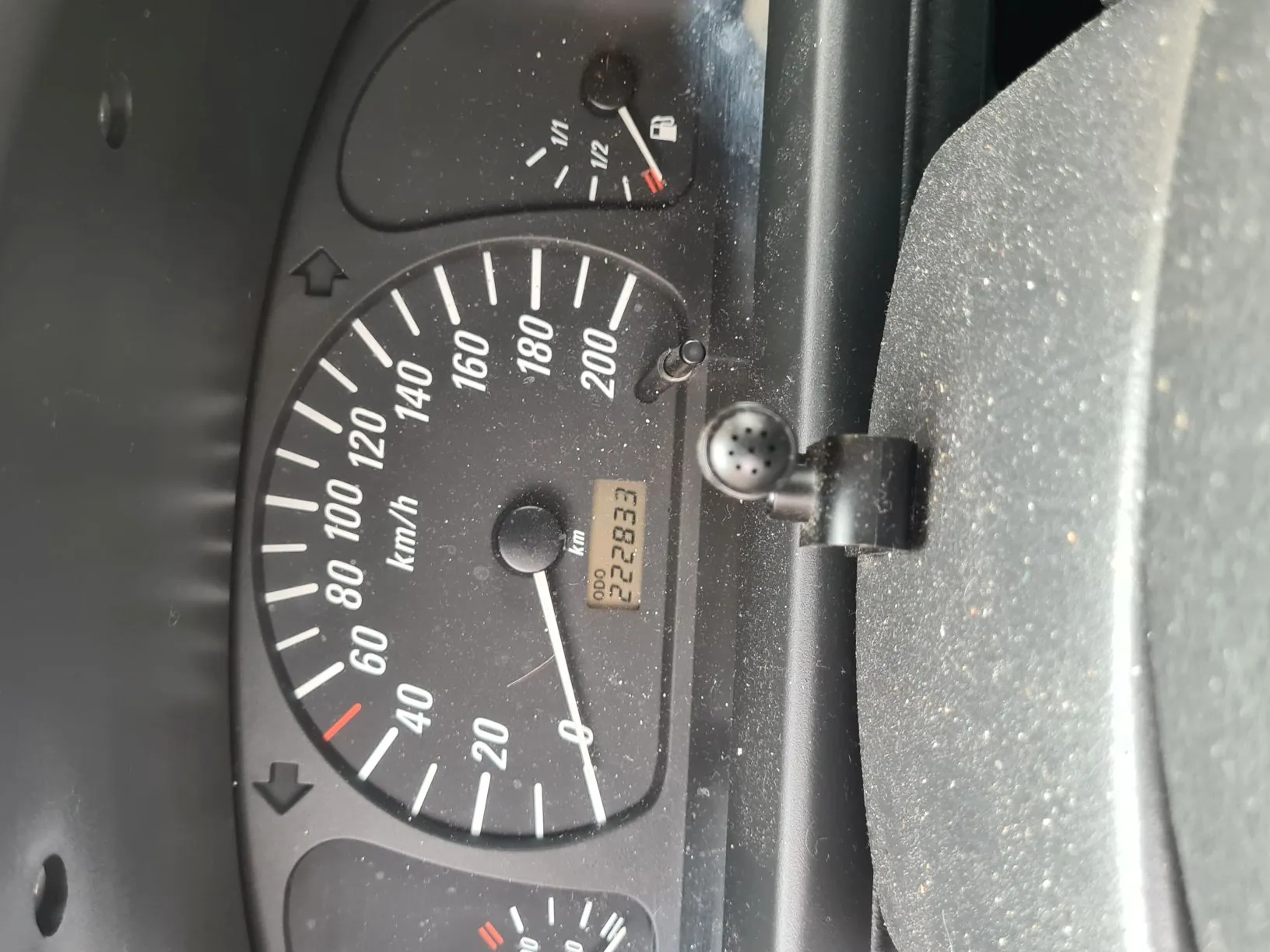 Zbliżenie na deski rozdzielcze samochodu. W centrum znajduje się prędkościomierz z zakresem do 200 km/h, wskazówka na 0 km/h. Obok prędkościomierza wyświetlacz przebiegu z napisem "ODO" i wartością 222833 km. Powyżej prędkościomierza wskaźnik paliwa z podziałką 1/1, 1/2 i ikoną zbiornika, wskazówka na poziomie około 1/2. Wszystkie elementy mają czarny tło z białymi znakami i cyframi. Na powierzchni deski rozdzielczej widoczny jest kurz.
