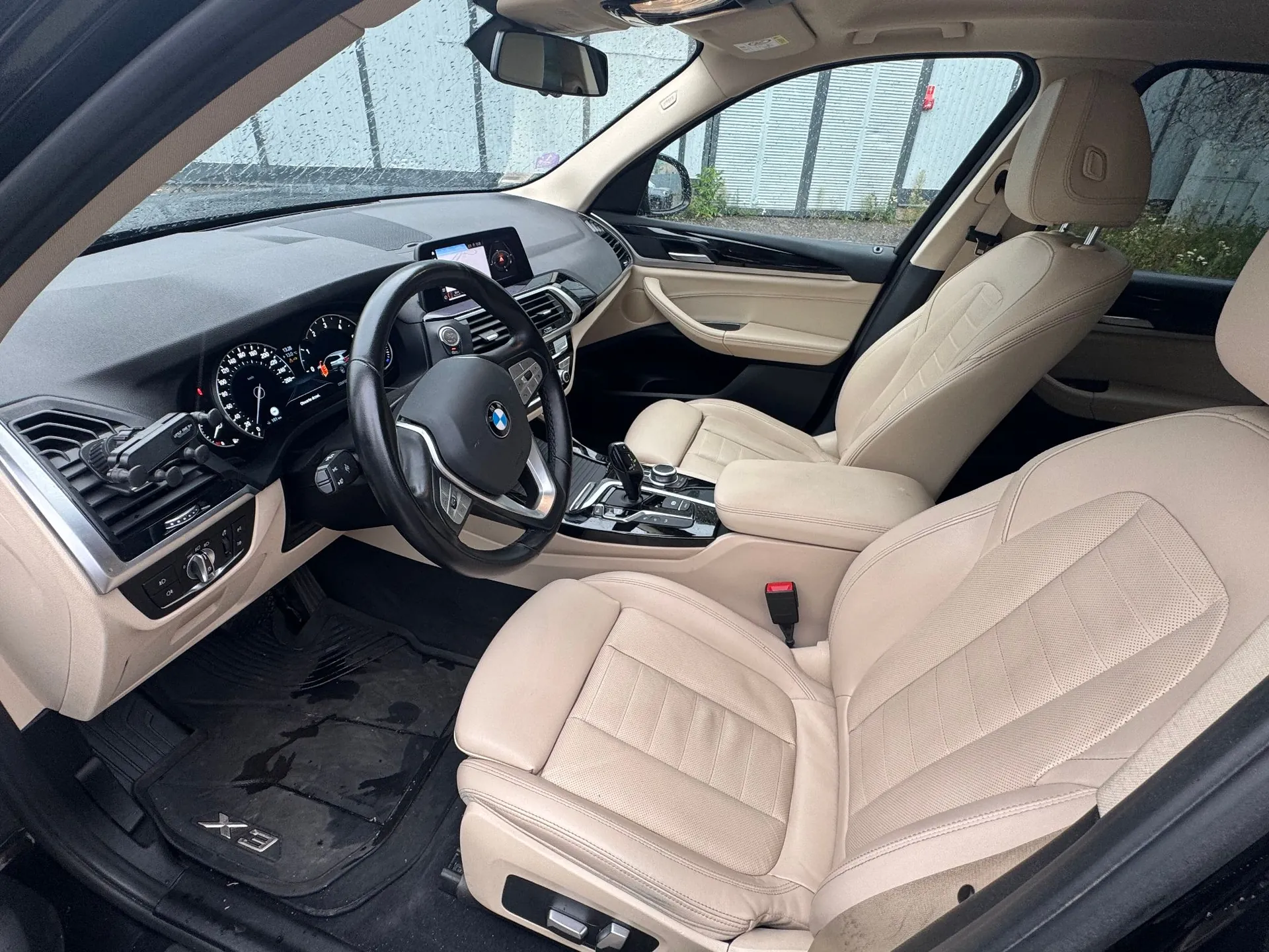 Wnętrze samochodu marki BMW z beżową, skórzaną tapicerką. Widoczna jest kierownica, deska rozdzielcza z ekranem multimedialnym, zegary analogowe oraz dwa przednie fotele. Na dywaniku kierowcy widnieje napis 'X3'.