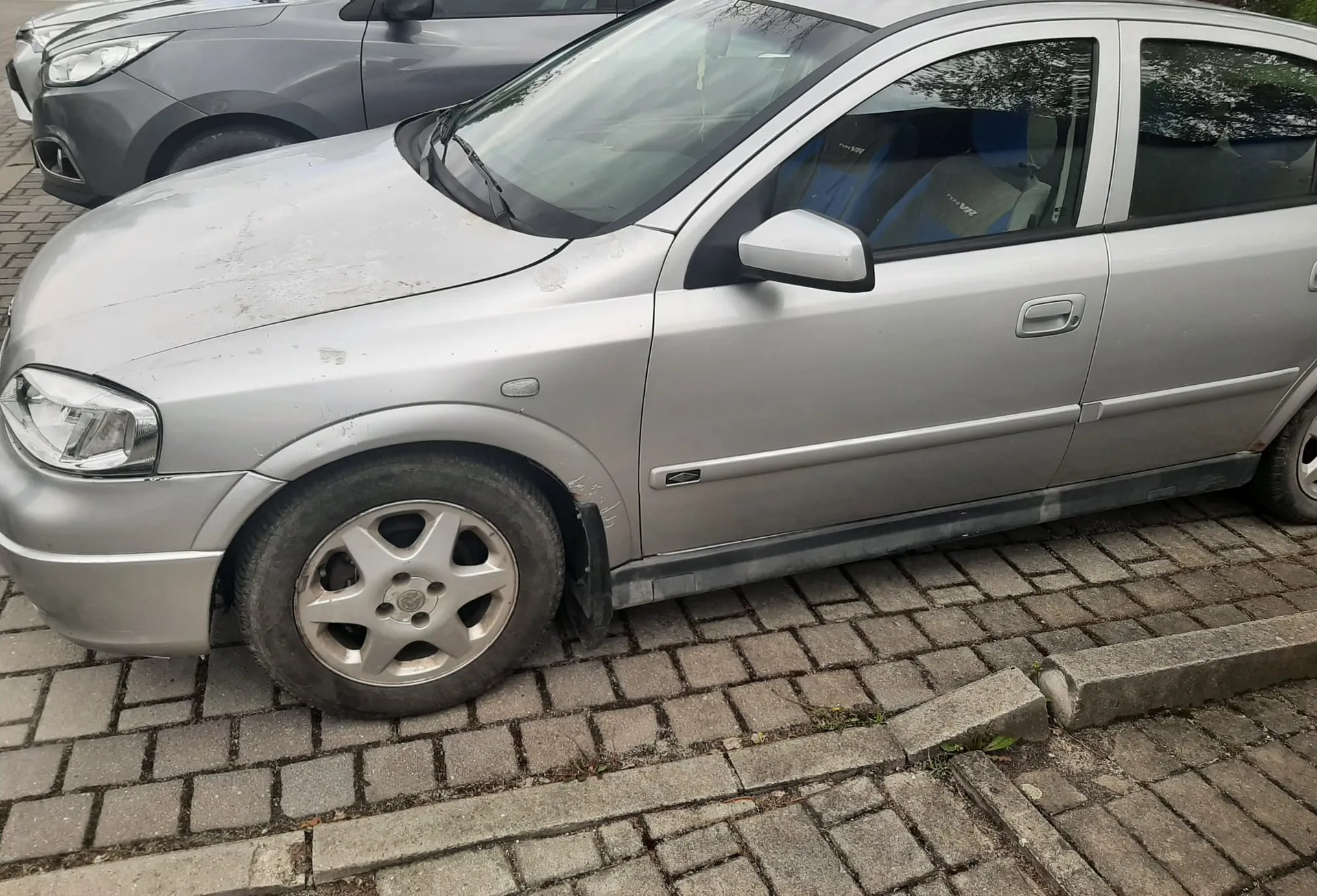 Srebrny samochód osobowy zaparkowany na brukowanej powierzchni. Pojazd ma widoczne zabrudzenia na maskie i kołach, srebrne felgi z pięcioma promieniami oraz niebieskie fotele wewnątrz. W tle częściowo widoczny szary samochód. Na pierwszym planie brukowa nawierzchnia z betonowym krawężnikiem, na którym rosną pojedyncze zioła.