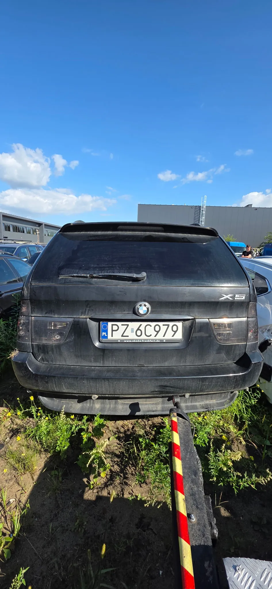 Tylny widok czarnego samochodu BMW X5 z polską tablicą rejestracyjną PZ 6C979. Pojazd jest brudny i znajduje się na terenie przypominającym złomowisko lub parking z trawą i roślinnością wokół. Samochód jest podniesiony na urządzeniu transportowym, z którego widoczny jest fragment czarnej ramy z czerwono-żółtymi pasami ostrzegawczymi.