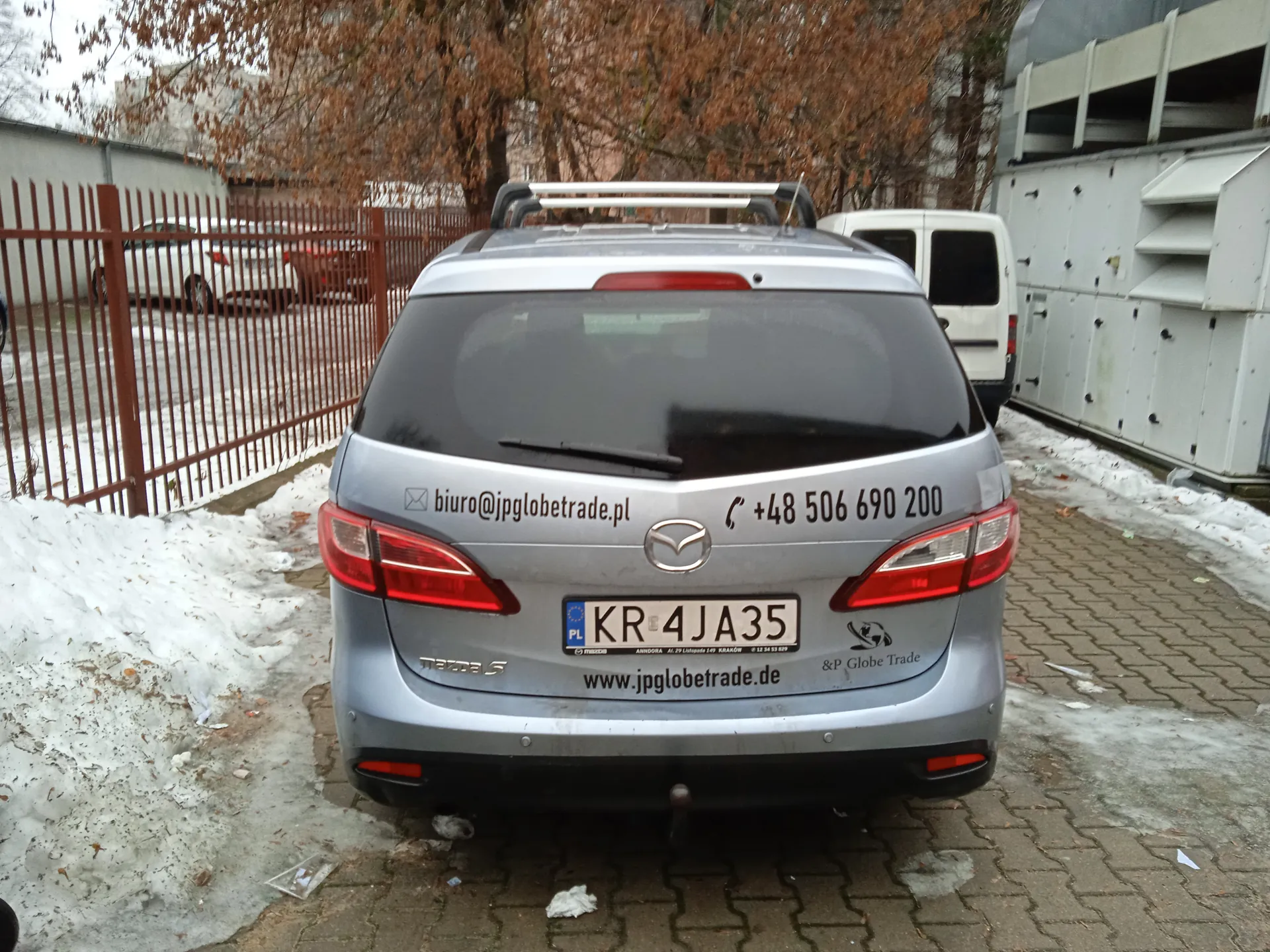 Tył srebrnego samochodu Mazda 5 z tablicą rejestracyjną KR 4JA35 (PL). Na bagażniku widoczne są napisy: 'biuro@jpglobetrade.pl', '+48 506 690 200', 'www.jpglobetrade.de' oraz logo '&P Globe Trade'. Samochód zaparkowany na brukowanej powierzchni z resztkami śniegu po bokach. Po lewej stronie czerwone metalowe ogrodzenie, za którym widać biały i czerwony samochód. W tle drzewa z brązowymi liśćmi, biały van oraz przemysłowe budynki z metalowymi konstrukcjami. Na ziemi rozrzucone są drobne śmieci.