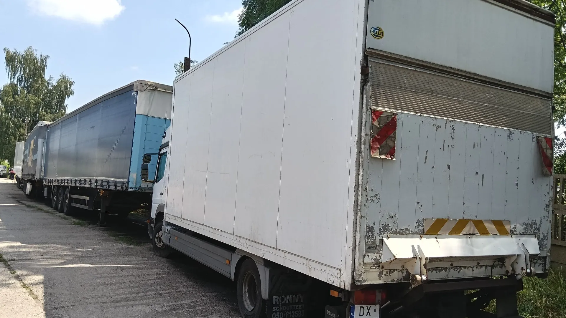 Zdjęcie przedstawiające dwa pojazdy transportowe zaparkowane obok siebie na utwardzonej drodze. Na pierwszym planie znajduje się biały samochód ciężarowy z zabudową furgonową. Na tylnej klapie widoczne są czerwono-białe pasy odblaskowe oraz biały zderzak. Na dolnej części nadbudowy znajduje się napis: 'RONNY SCHOUTTEET 050/893583'. Za nim zaparkowana jest niebieska naczepa ciężarowa z plandeką. W tle widoczne są zielone drzewa i jasne niebo z lekkimi chmurami.