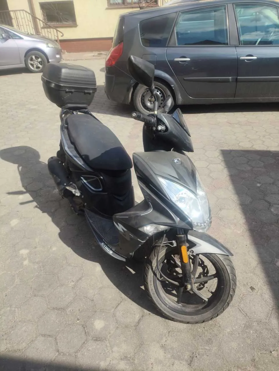 Czarny skuter marki KYMCO z czarnym bagażnikiem na tylnej części, zaparkowany na brukowanej powierzchni. W tle widoczne są dwa samochody: ciemnoszary minivan i srebrny samochód osobowy. Za nimi znajduje się budynek z oknami i schodami. Na skuterze widoczny jest napis 'KYMCO' na boku. Oświetlenie słoneczne rzuca cienie na bruk.