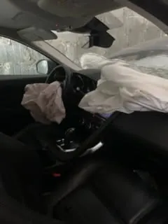 Wnętrze samochodu z rozwiniętymi poduszkami powietrznymi po stronie kierowcy i pasażera. Widoczne czarne siedzenia, kierownica, deska rozdzielcza oraz dźwignia zmiany biegów. Na zewnątrz przez przednią szybę widać kamienną ścianę. Brak czytelnego tekstu.