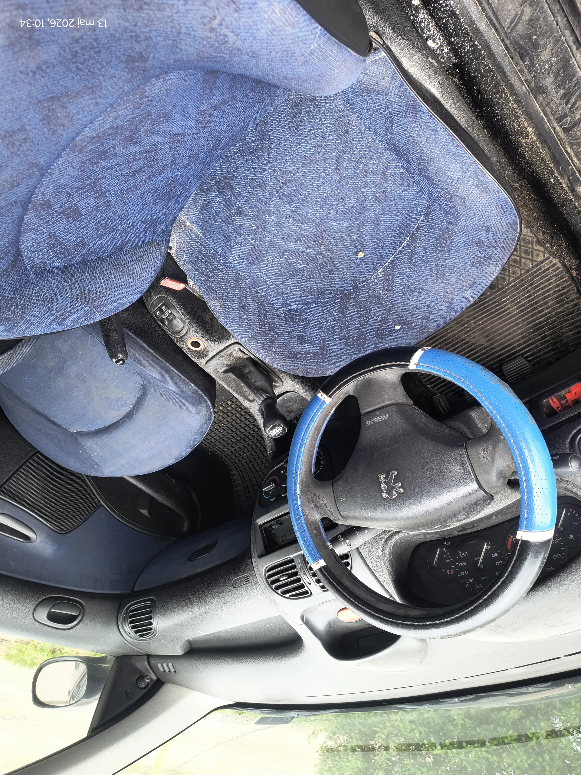 Wnętrze samochodu z widocznym kierownicą marki Peugeot z niebieską obwódką, niebieskimi siedzeniami tkaninowymi, deską rozdzielczą z wentylacjami i wskaźnikami. Na siedzeniach widoczne są zabrudzenia. Po prawej stronie obrazu znajduje się pionowy tekst: 13 maj 2026, 10:34. W tle przez okno widać zieloną roślinność.