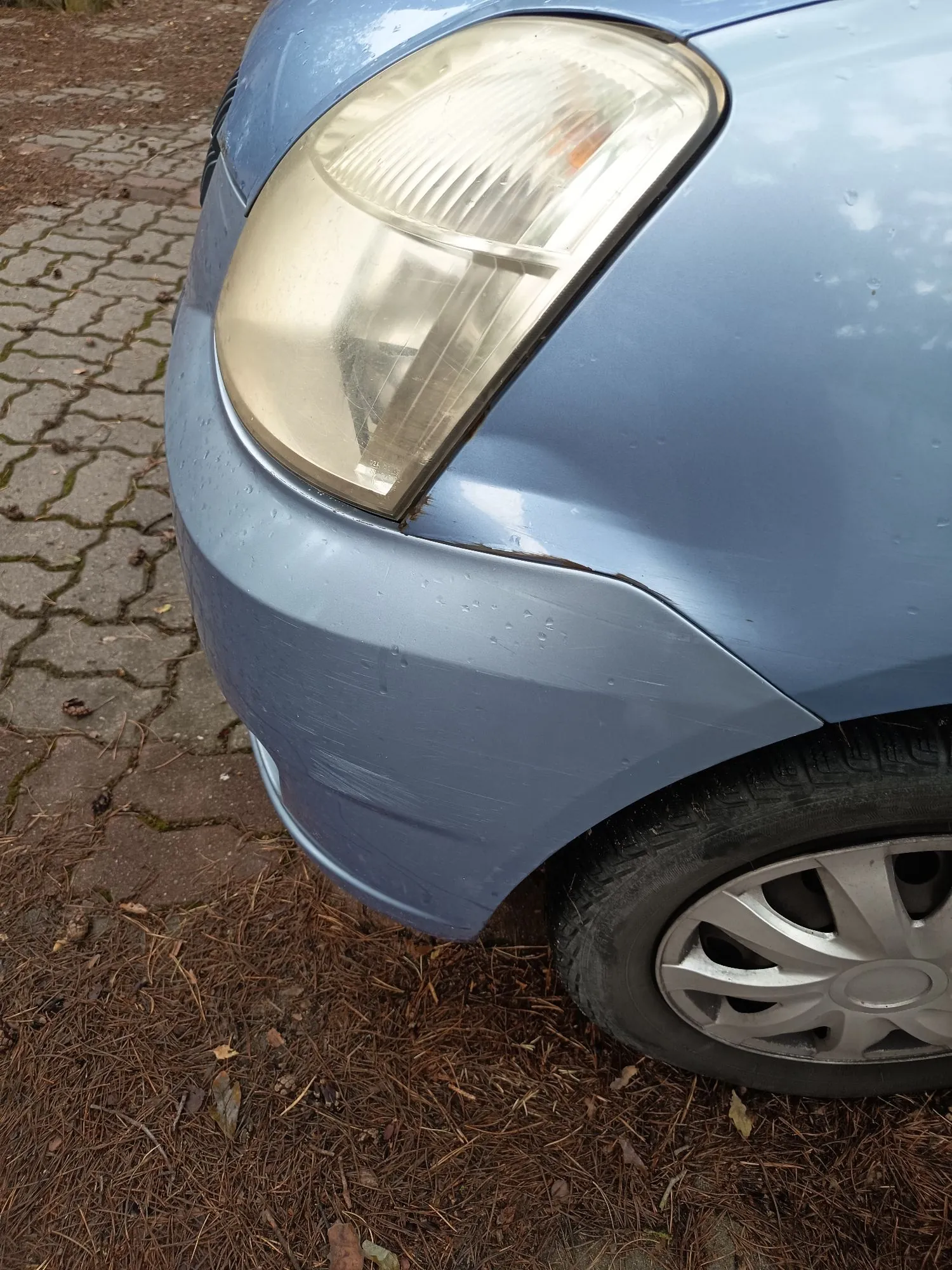 Zbliżenie na przedni prawy narożnik jasnoniebieskiego samochodu. Widoczna jest zmatowiała lampa przednia oraz wgniecenie i zarysowania na zderzaku tuż pod reflektorem. W dolnej części obrazu widoczny jest fragment koła z felgą aluminiową, a samochód stoi na podłożu z kostki brukowej i igliwia.