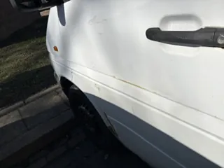 Biały samochód widziany z boku z czarną klamką drzwi. Na boku pojazdu widoczna jest brudna plama lub zadrapanie. Część przedniego koła oraz chodnik z trawnikiem w tle.