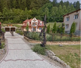 Widok na posesję z domem jednorodzinnym o czerwonym dachu i jasnych ścianach. Przed domem znajduje się ozdobna, biała ścieżka z kostki brukowej z czerwonymi akcentami, prowadząca w stronę budynku. Po prawej stronie ścieżki znajduje się ogród z niskim ogrodzeniem i zielenią, a w tle gęsty, zielony las na wzgórzu. Po prawej stronie widoczny jest fragment innego, białego budynku.