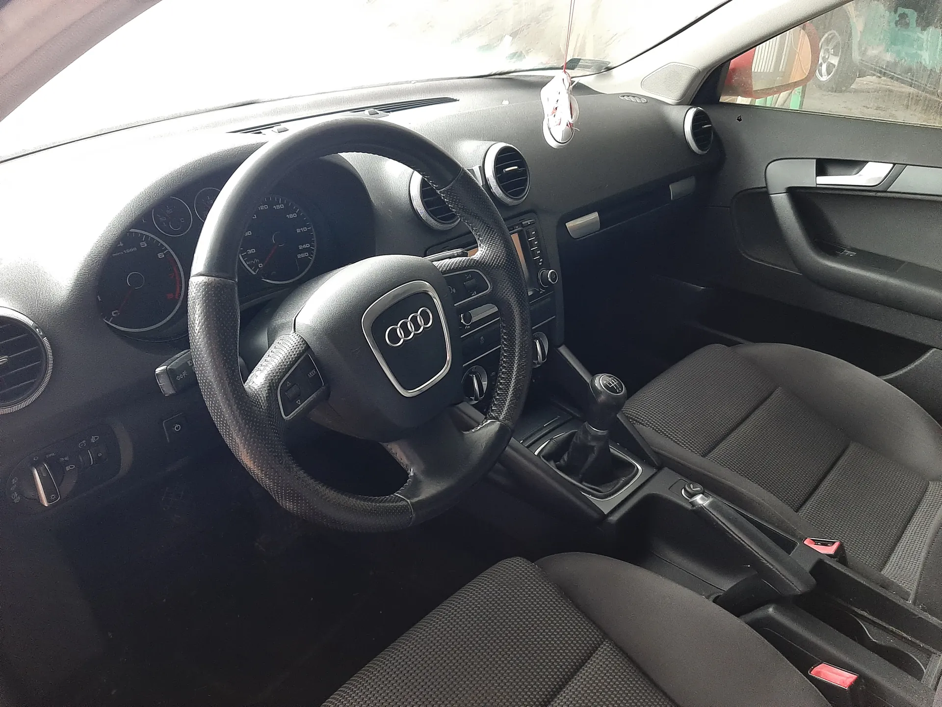 Wnętrze samochodu marki Audi z widocznym kierownicą z logo czterech pierścieni, czarnymi siedzeniami z teksturą, deską rozdzielczą z licznikami prędkości i obrotomierzem, przyciskami sterującymi, klimatyzacją z okrągłymi wydmuchami, dźwignią zmiany biegów oraz czerwonymi zapięciami pasów bezpieczeństwa. Na lusterku wstecznym wiszą ozdoby. Widoczne są również drzwi z uchwytem i oknem.